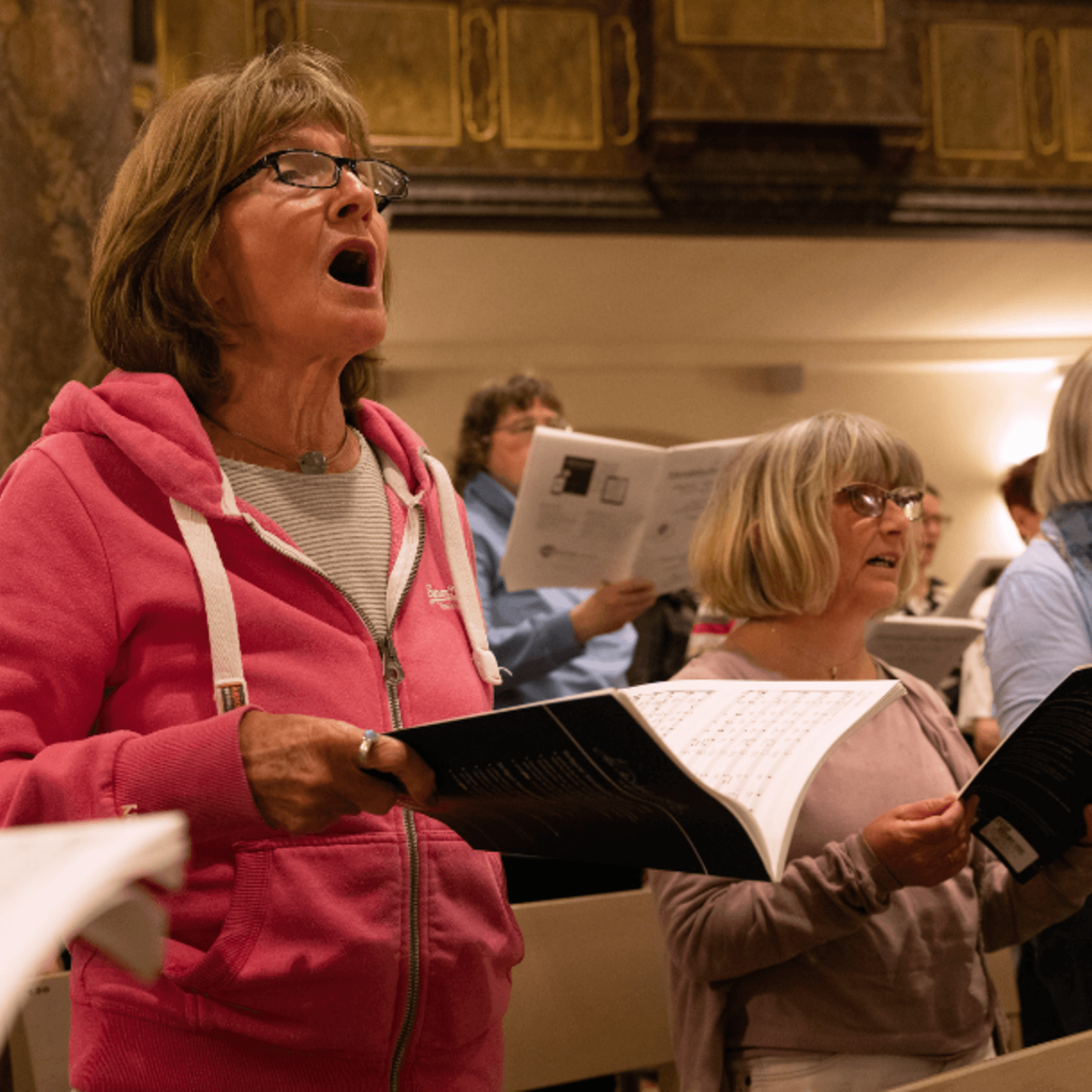 Eine Frau im Vordergrund singt leidenschaftlich bei der Chorprobe. Weitere Menschen mit Notenheft beteiligen sich rechts von ihr ebenfalls im Chor.