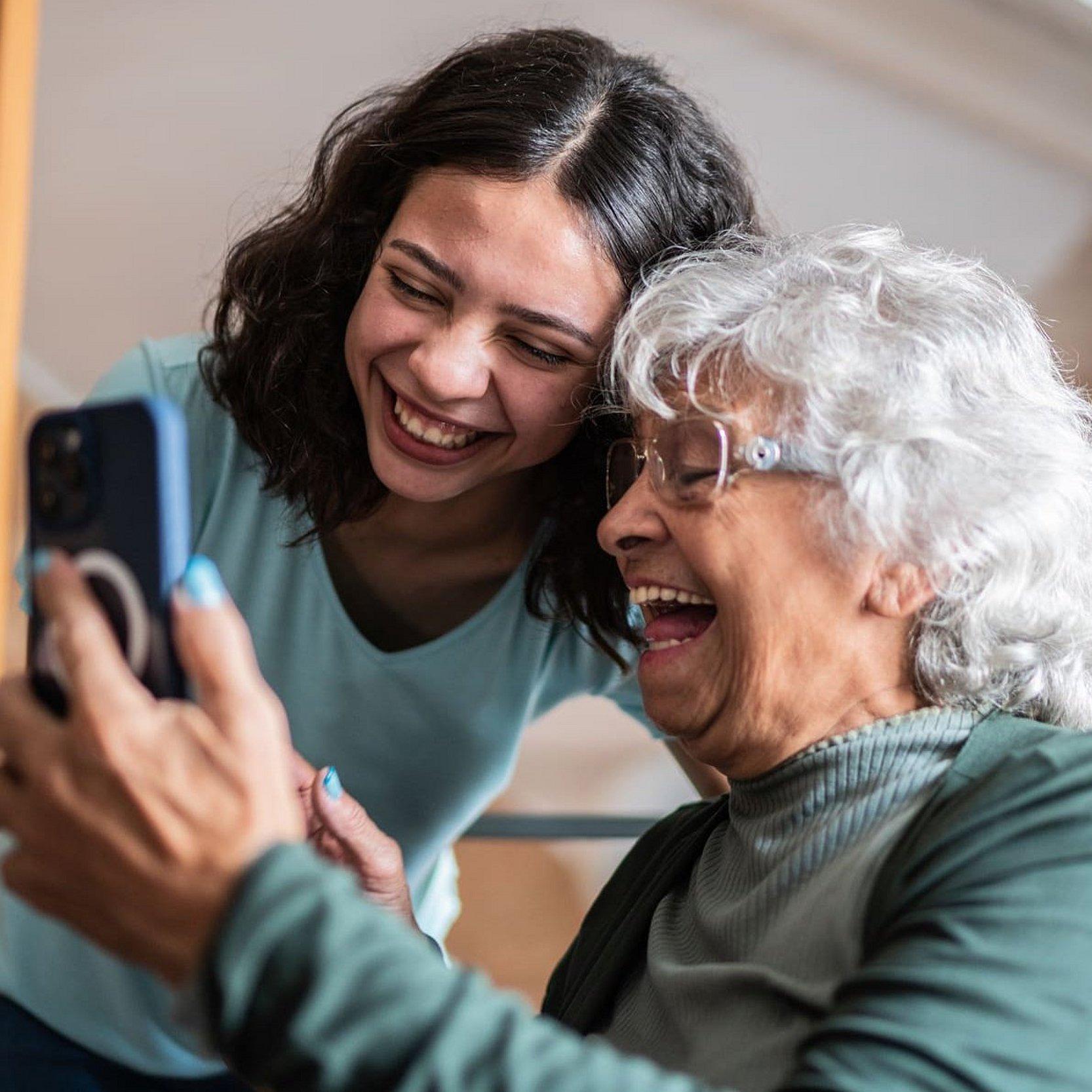 Eine ältere Frau sitzt und hält ein Smartphone in der Hand, eine junge Frau beugt sich über sie und schaut auch in das Smartphone, beide lächeln und sehen sehr freundlich aus.