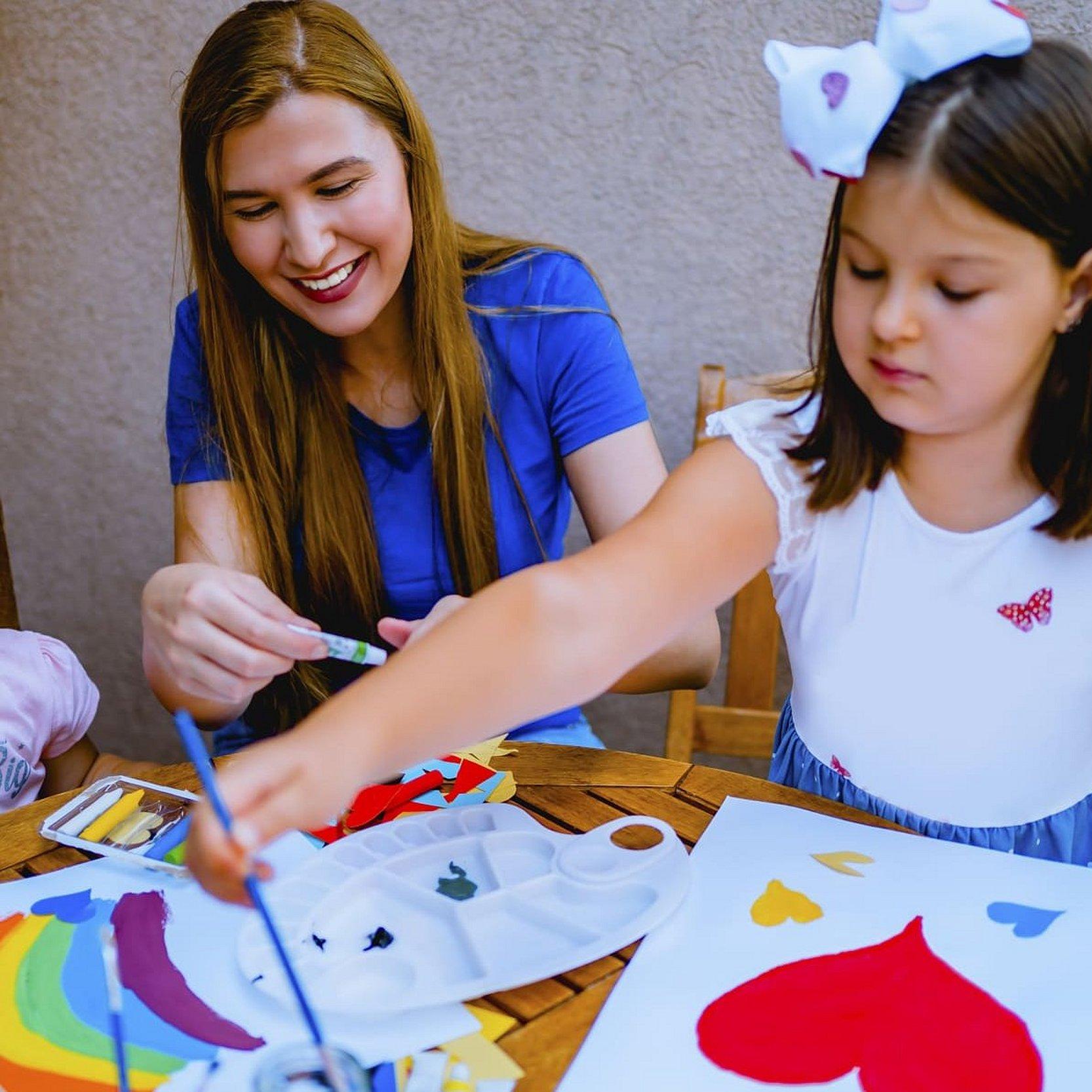 Eine Mutter sitzt in der Mitte am Tisch, rechts und links jeweils ein Mädchen, ihre Töchter, die Bilder mit Wasserfarbe malen. Auf einem Bild ist ein rotes Herz zu sehen, auf dem anderen Bild ein Regenbogen in vielen Farbein, alle sehen glücklich aus.