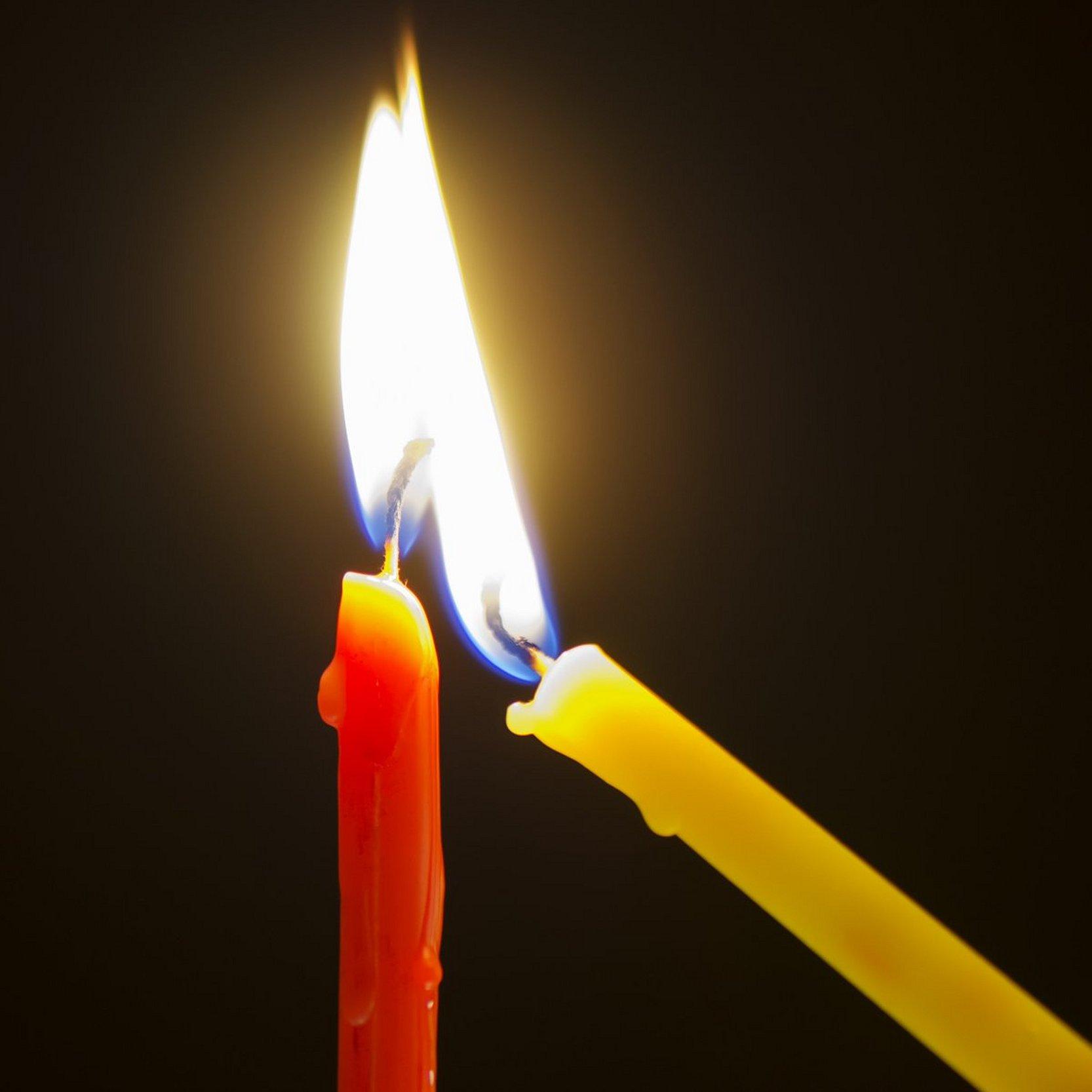 Eine gelbe schmale Kerze wird an einer roten schmalen Kerze angezündet.