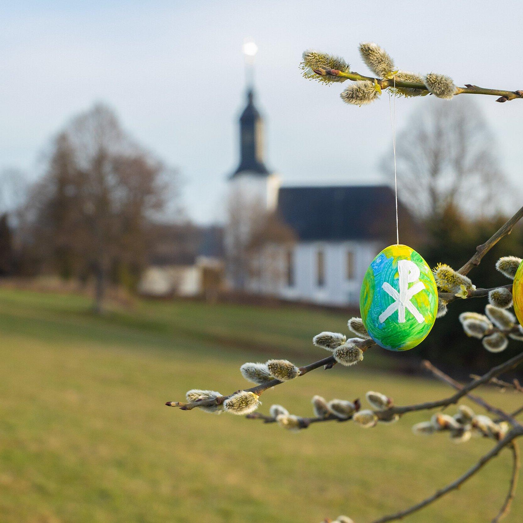 Bunt bemalte Ostereier hängen an einem Baum. Im Hintergrund ist eine Dorfkirche zu sehen.