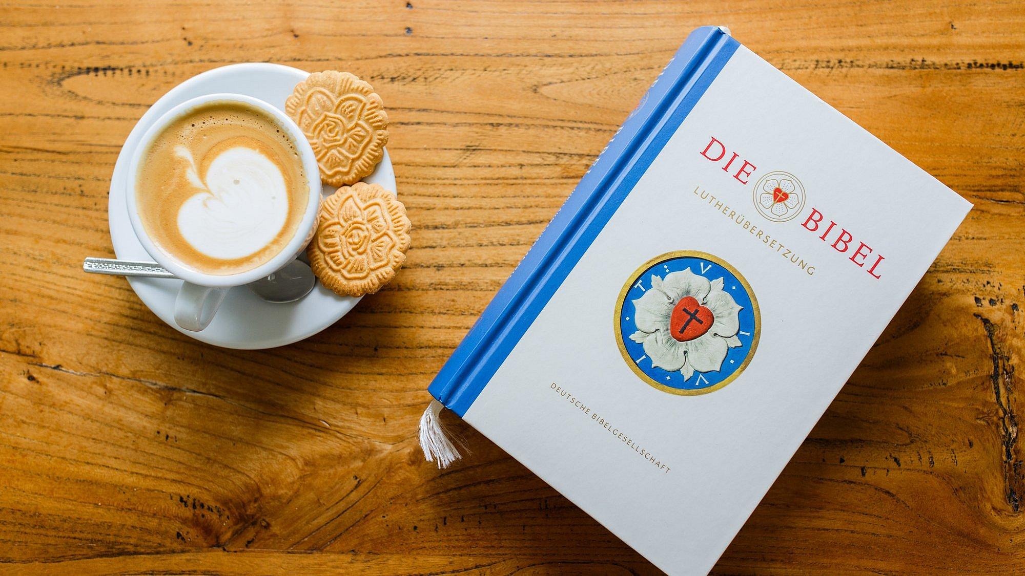 Lutherbibel neben einer Kaffeetasse mit Milchschaum in Apfelform und Keksen in Form der Lutherrose