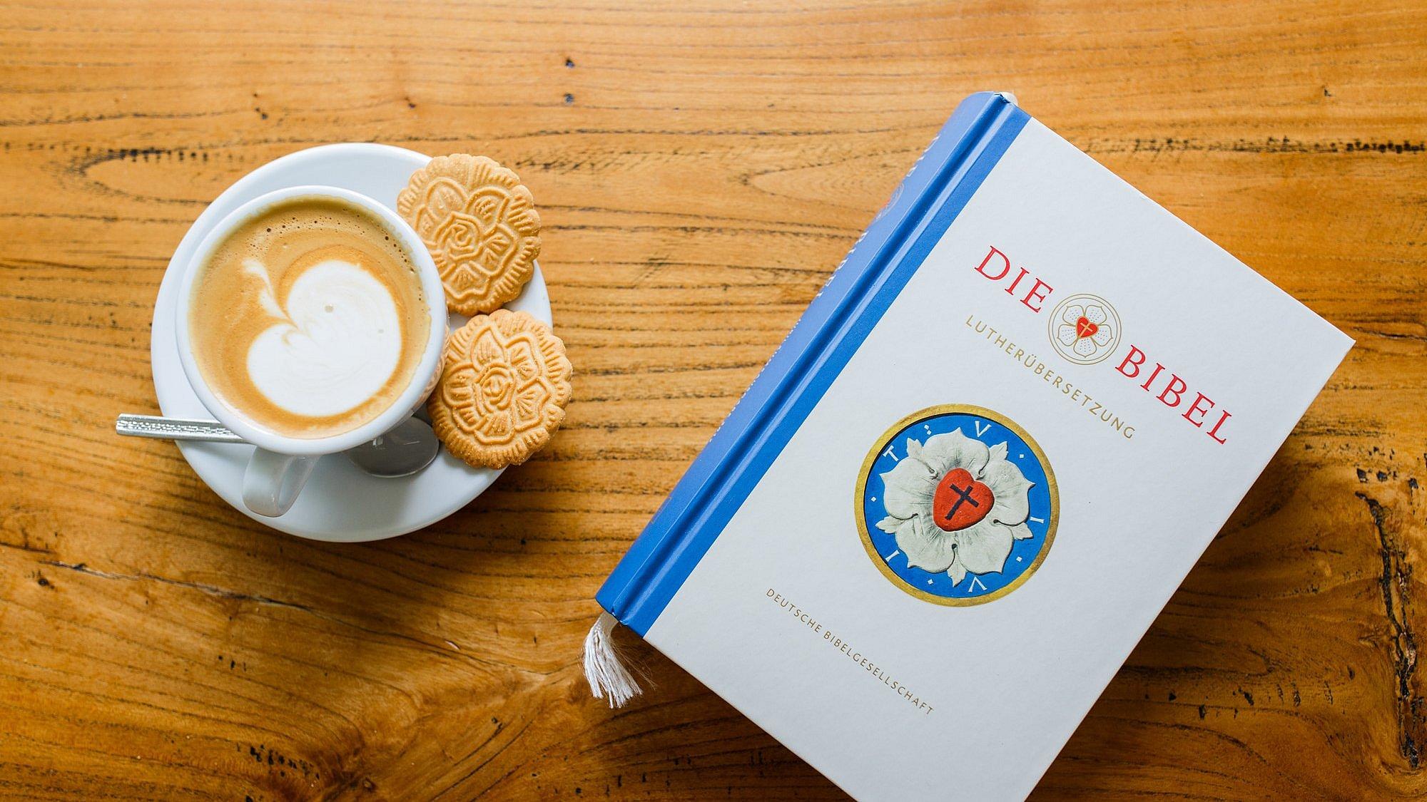 Bibel Lutherbibel neben einer Kaffeetasse mit Milchschaum in Apfelform und Keksen in Form der Lutherrose