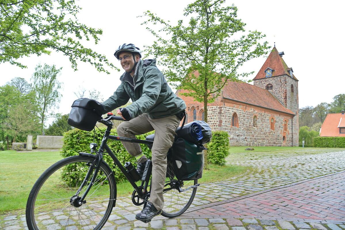 Ein fröhlicher Radfahrer fährt von rechts nach links durch das Bild. Im Hintergrund ist eine gepflegte grüne Landschaft um eine Kirche zu sehen.