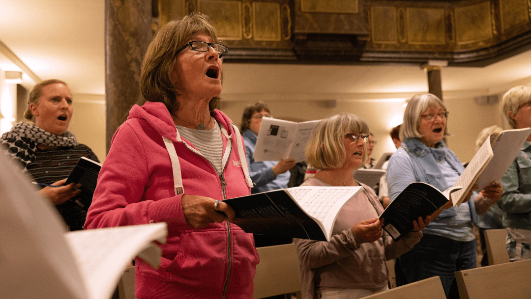 Eine Frau im Vordergrund singt leidenschaftlich bei der Chorprobe. Weitere Menschen mit Notenheft beteiligen sich rechts von ihr ebenfalls im Chor.