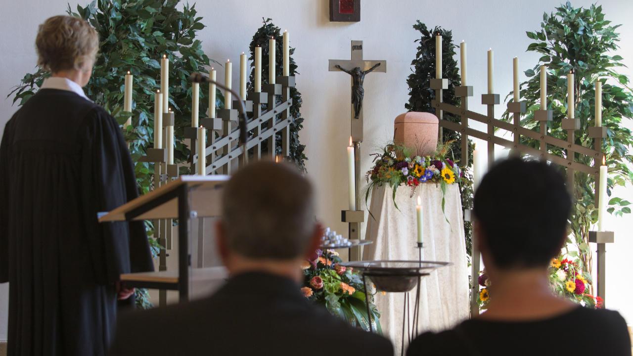 eine Urne auf einem Sockel, Kerzen und ein Kreuz in der Trauerhalle auf dem Friedhof, zwei Trauernde von hinten, eine evangelische Pfarrerin, 