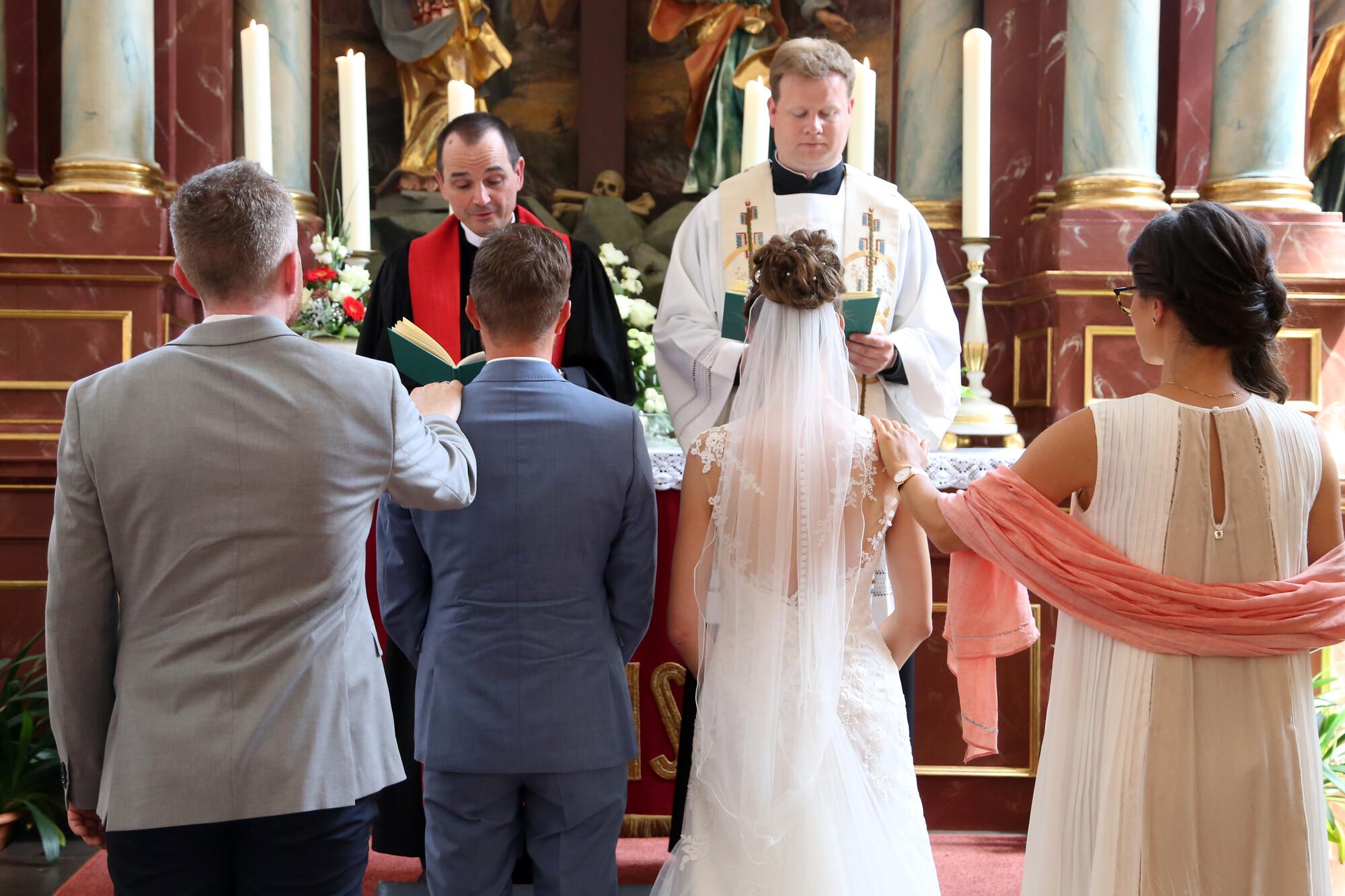 Ein Brautpaar mit Trauzeugen vor dem Altar. Vor dem Altar stehen ein katholischer und ein evangelischer Pfarrer