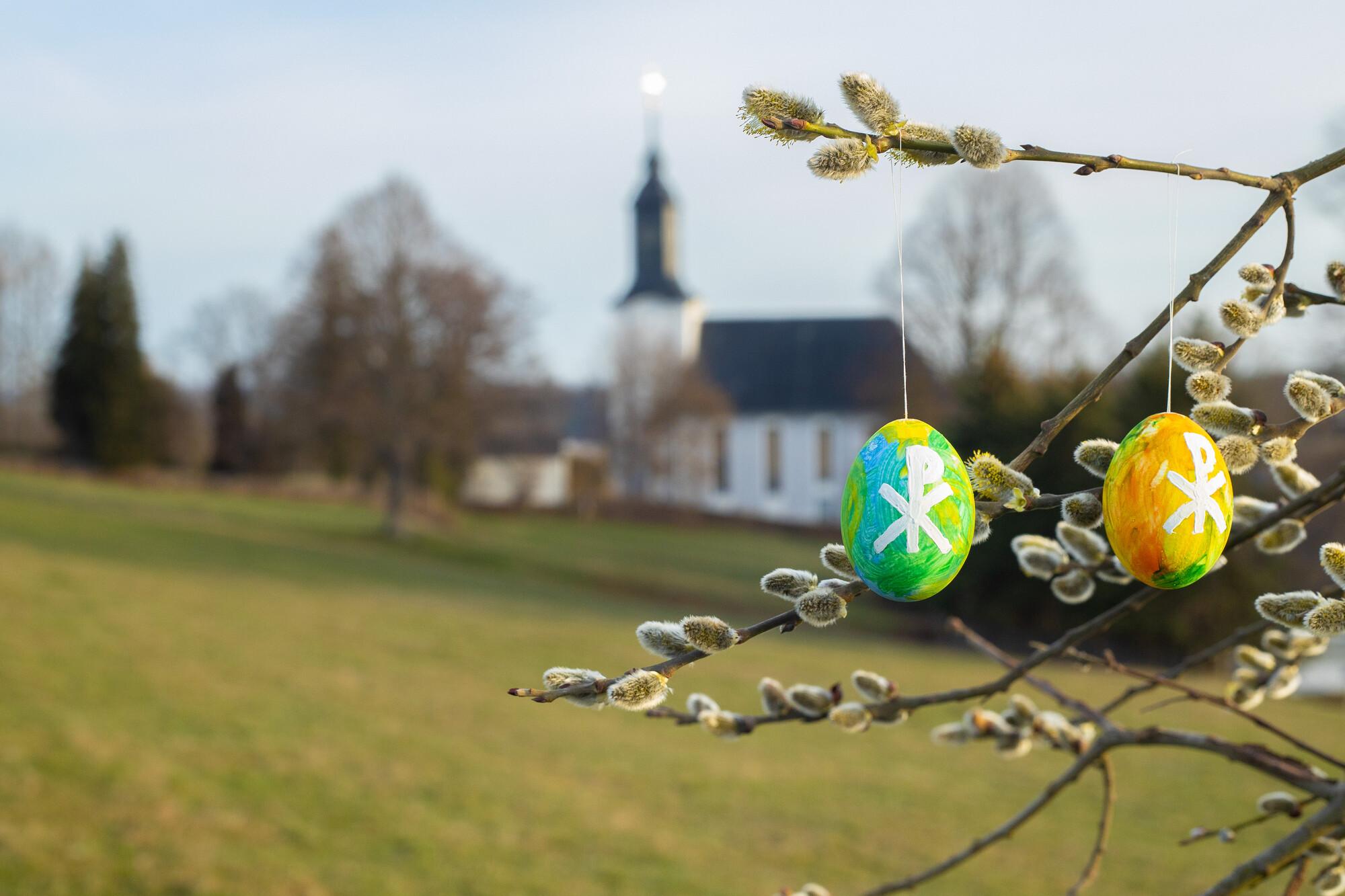 Bunt bemalte Ostereier hängen an einem Baum. Im Hintergrund ist eine Dorfkirche zu sehen.