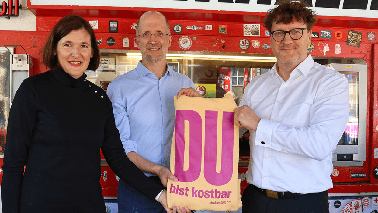 Drei Personen halten ein Plakat mit dem Kampagnenmotiv "DU bist kostbar"
