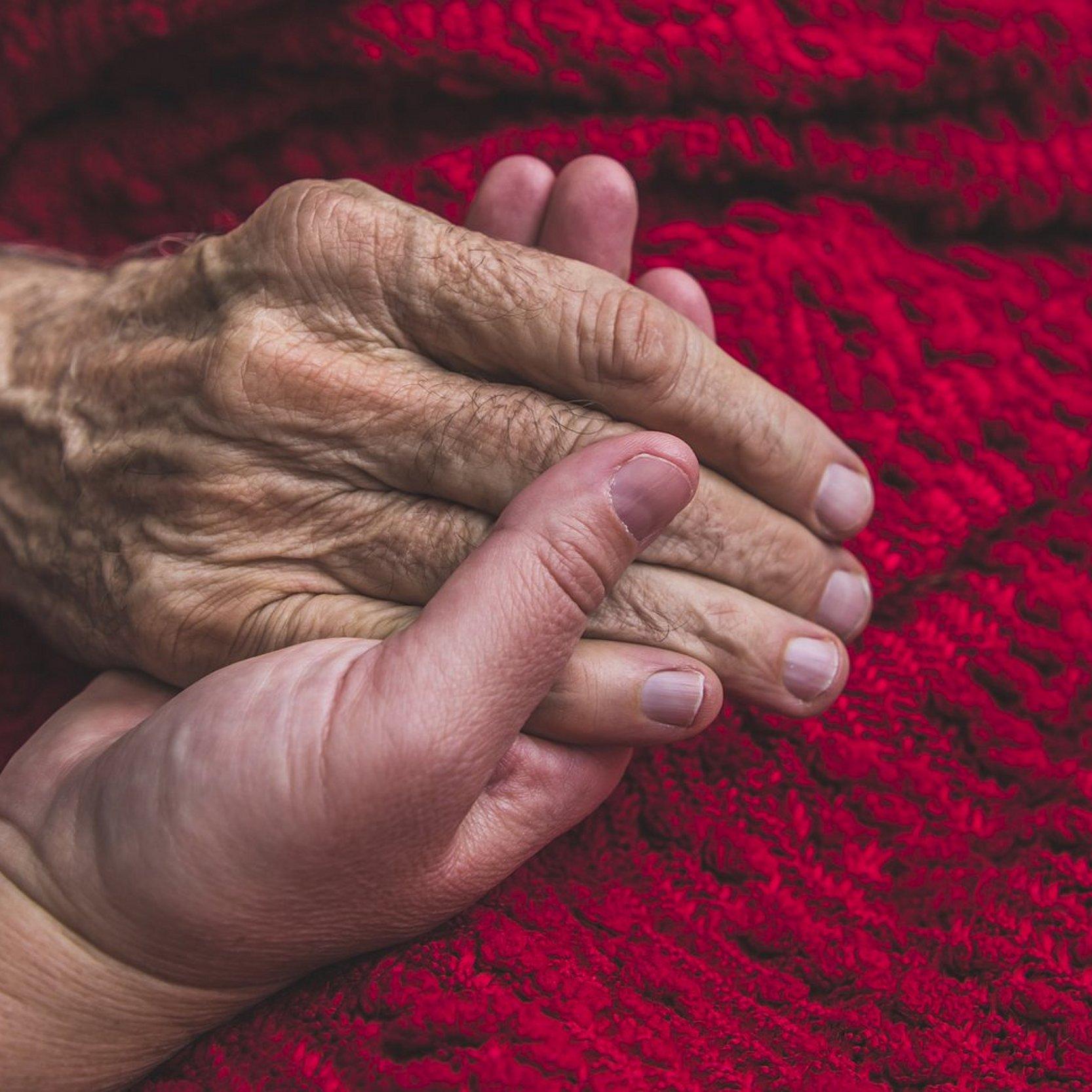 Eine junge Hand hält die alte Hand einer Sterbenden. Diese ist mit einer schönen, roten Decke zugedeckt.