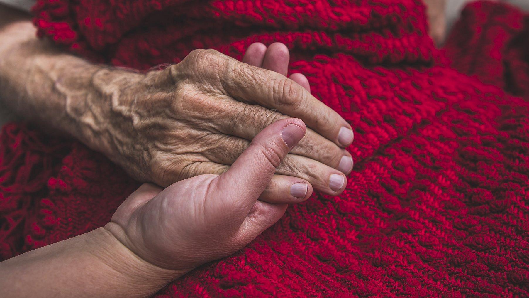 Eine junge Hand hält die alte Hand einer Sterbenden. Diese ist mit einer schönen, roten Decke zugedeckt.