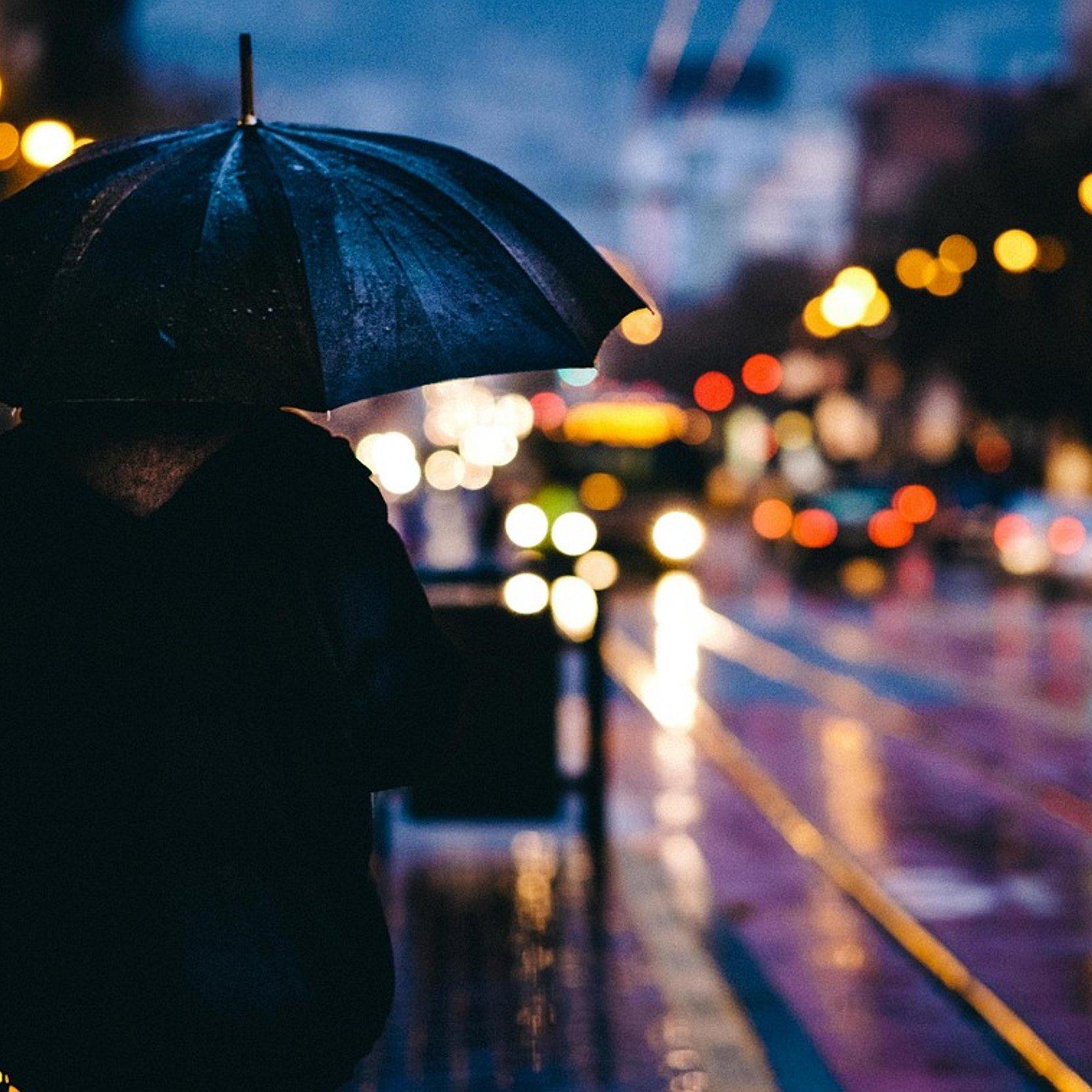 „Eine Person mit Regenschirm steht bei Regen an einer beleuchteten Straße in der Dämmerung. Das unscharfe Stadtlicht erzeugt eine nachdenkliche und ruhige Atmosphäre.“