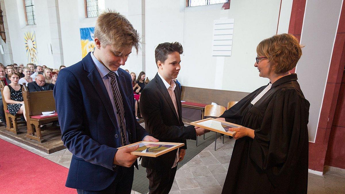 Zwei Konfirmanden stehen mit dunklen Anzügen vor der Pfarrerin im Talar. Die Pfarrerin überreicht den beiden den Segenspruch.