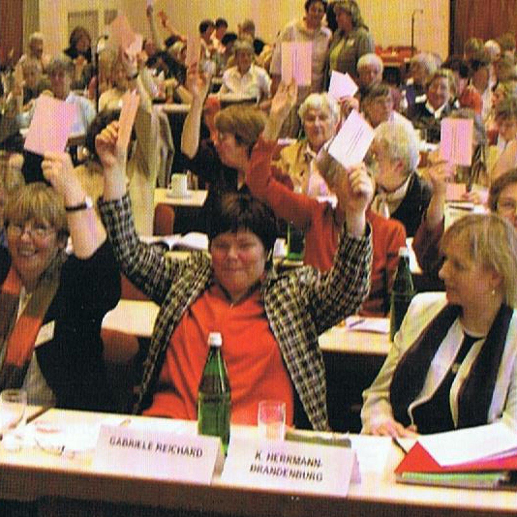 Ein großer Saal mit vielen Frauen, die an Tischen sitzen, wie auf einer Konferenz und mit erhobenden Händen abstimmen