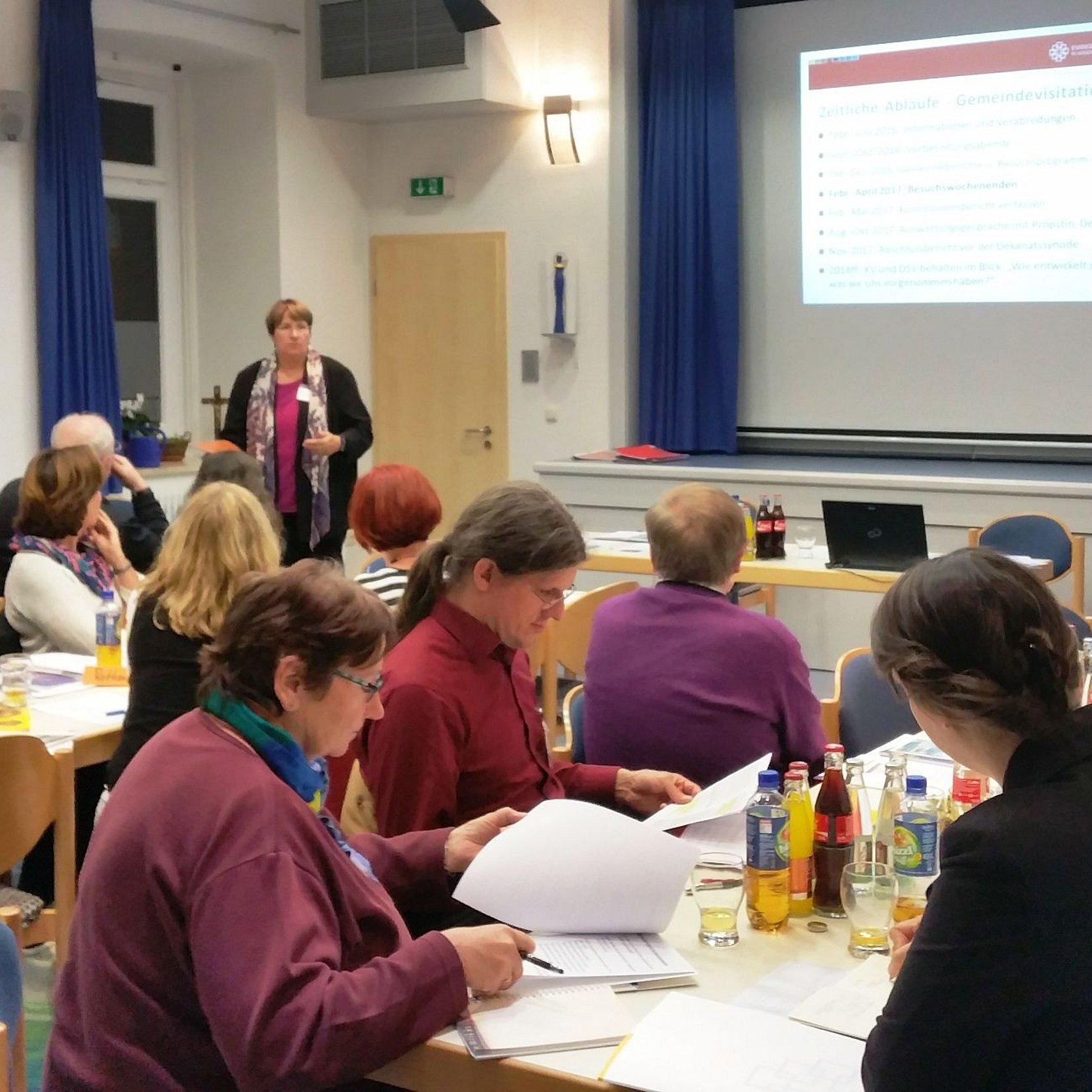 Menschen an Konferenztischen, im HIntergrund Präsentation auf Leinwand