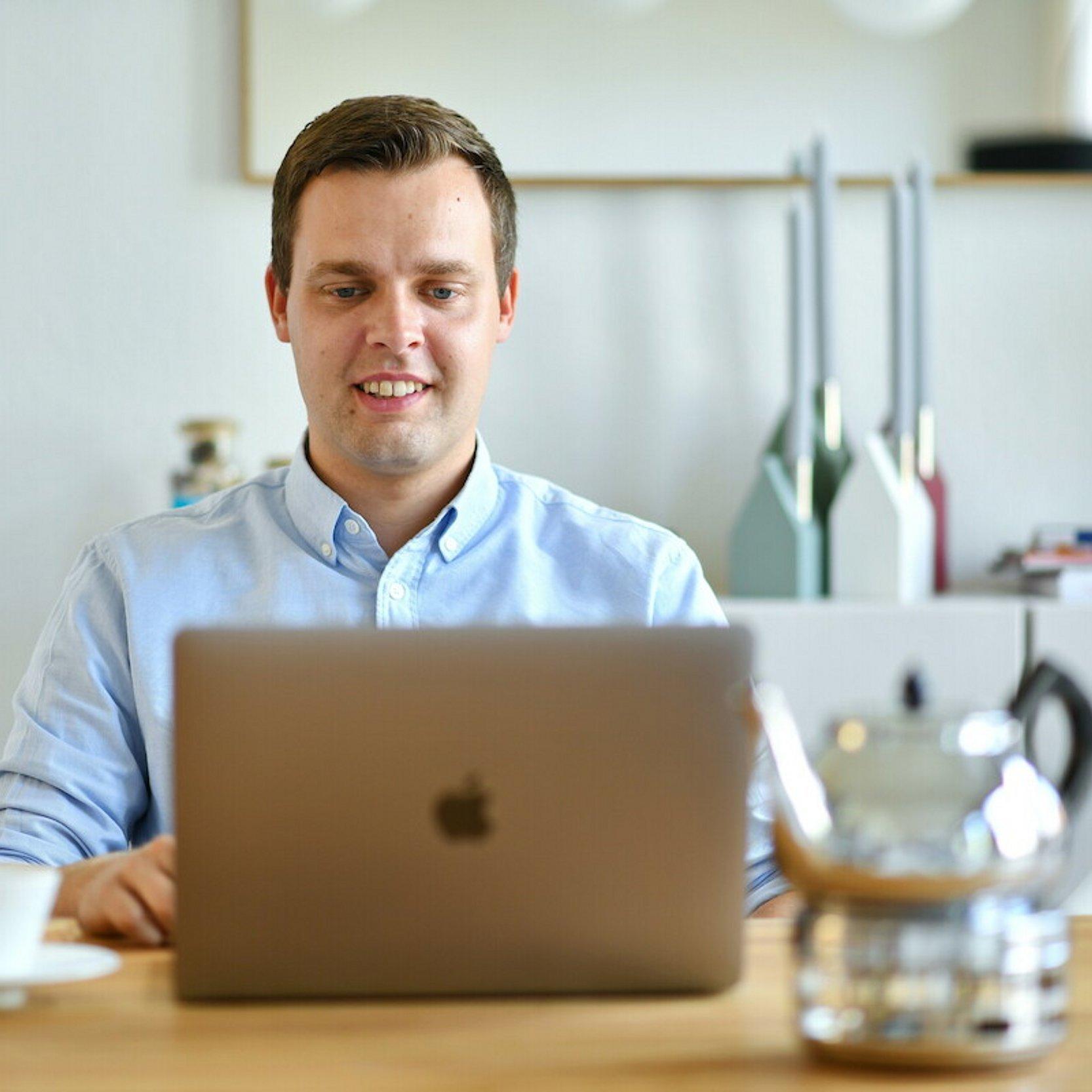 Ein Mann sitzt an einem gedeckten Küchentisch und schaut in den Monitor seines Notebooks. Im unscharfen Vordergrund stehen auf dem Tisch eine Teekanne, Kaffeetassen, Teller und Gläser. Im unscharfen Hintergrund ist eine Wand mit einem Spiegel zu erkennen, außerdem ein Kerzenständer und eine Tischlampe.