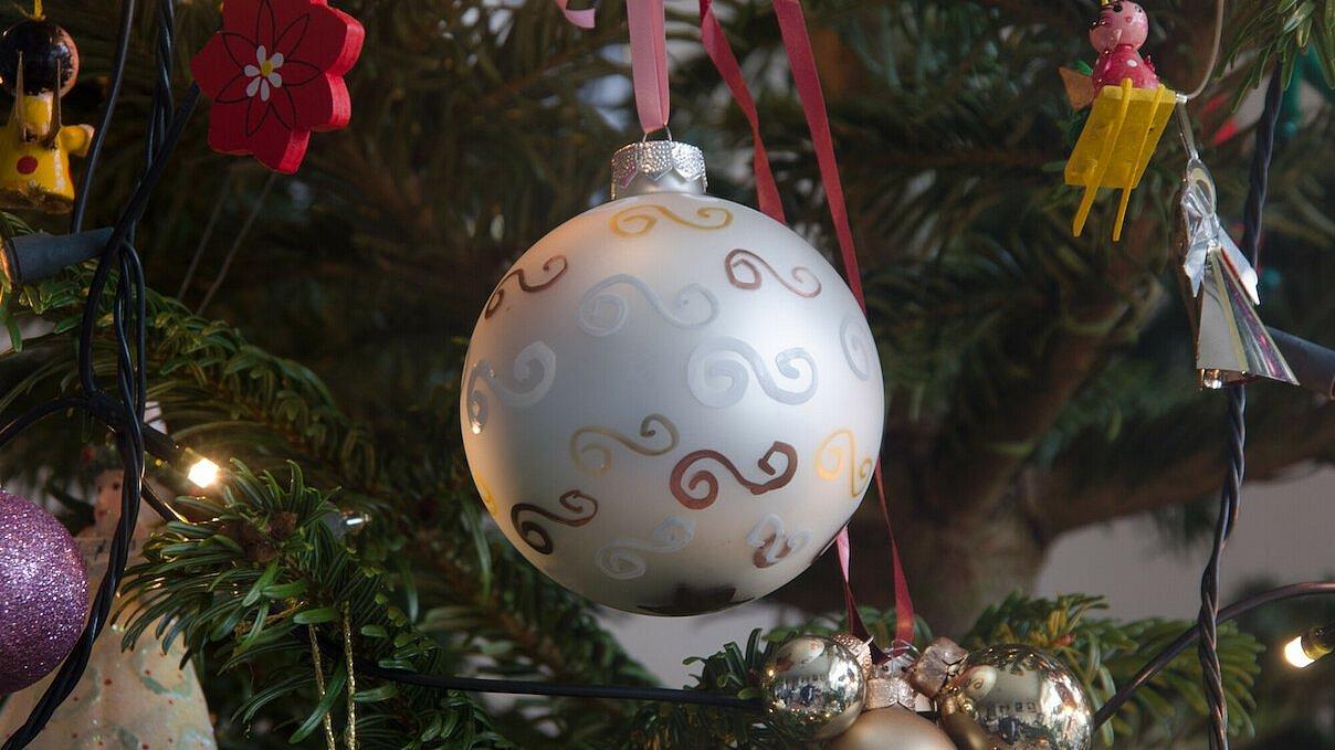 Eine silberfarbene Christbaumkugel vorne und weiterer Christbaumschmuck, Engel und Schleifen, die an den Zweigen eines Christbaumes hängen.