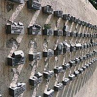 Zu sehen sind Gedenktafeln an der Mauer des jüdischen Friedhofs Battonstraße in Frankfurt/M für die aus der Stadt deportieren jüdischen Menschen. Seitlich an der Mauer befinden sich kleine Boxen, in welche Namen sowie Geburts- und Todesdaten eingraviert sind. Auf diesen Boxen liegen kleine Gedenksteine.