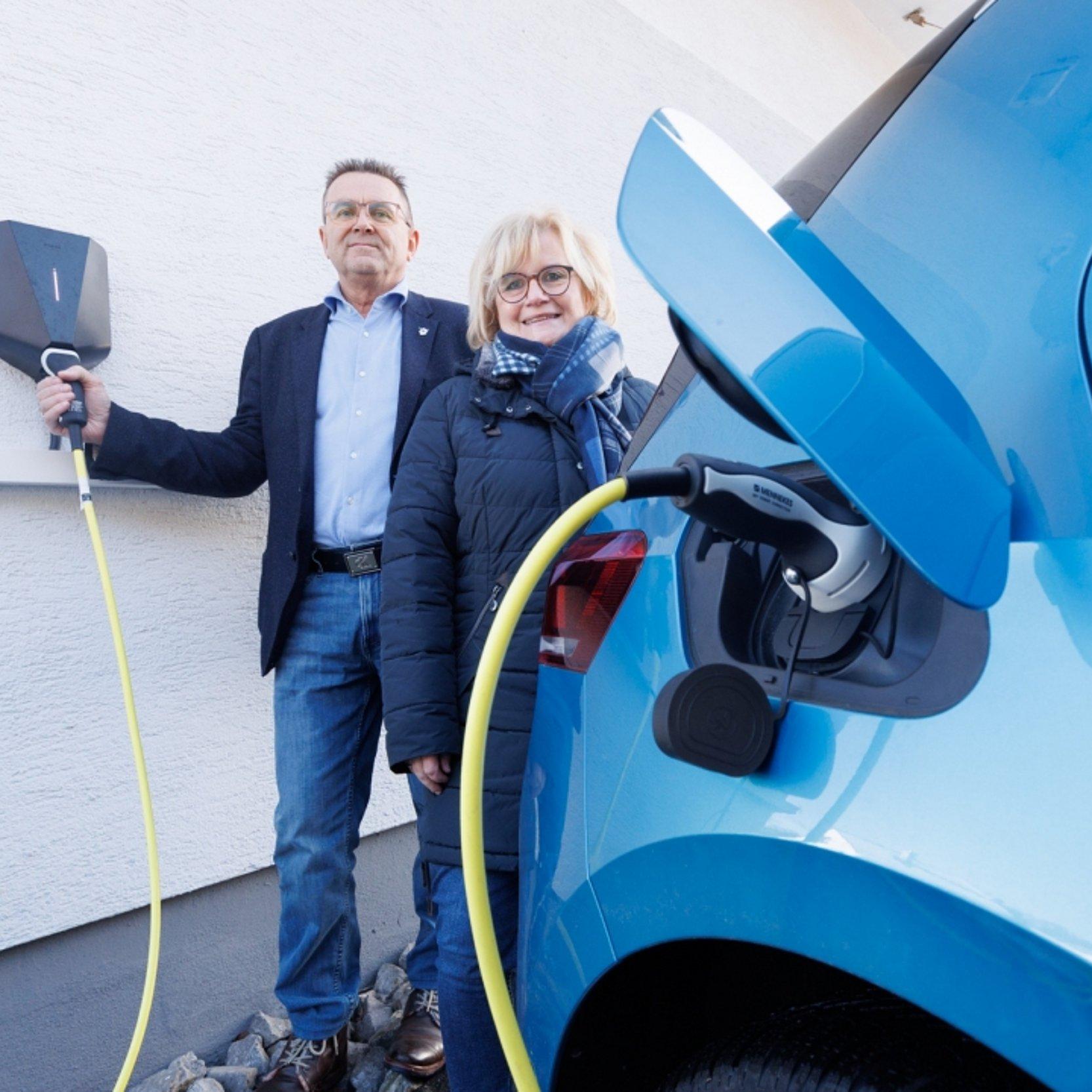 Ein Mann und eine Frau stehen neben einem blauen Elektroauto, das über eine Wallbox an der Hauswand geladen wird. Beide halten Ladekabel in der Hand und blicken in die Kamera.