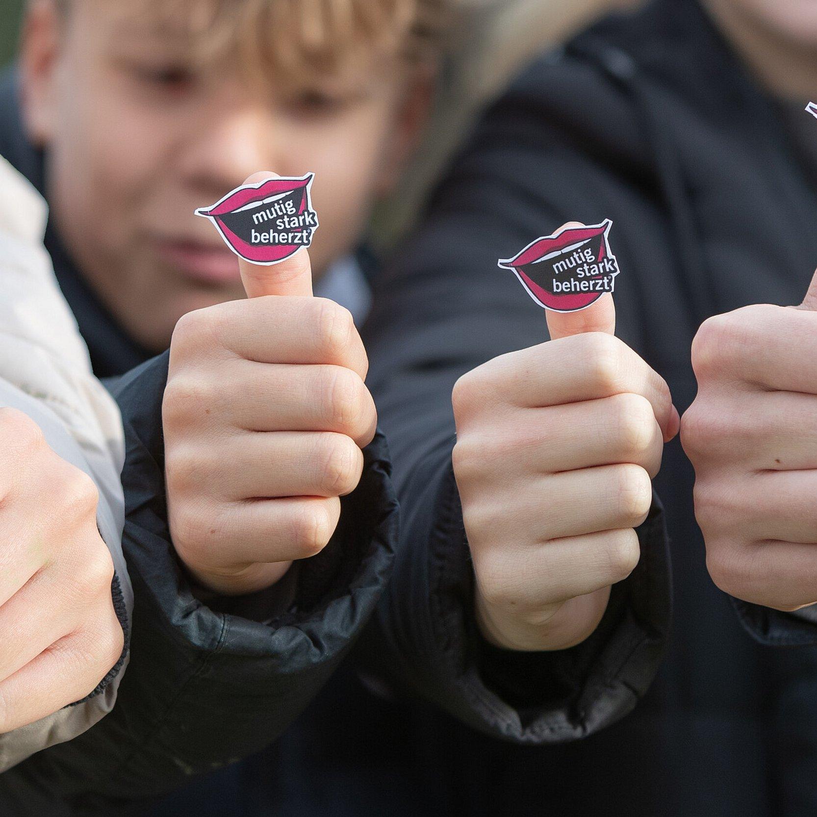 Zwei Konfirmanden und zwei Konfirmandinnen tragen das Logo des Kirchentages 2025 in Hannover (Offener Mund mit den Worten "mutig", "stark" und "beherzt") an ihrem Daumen.