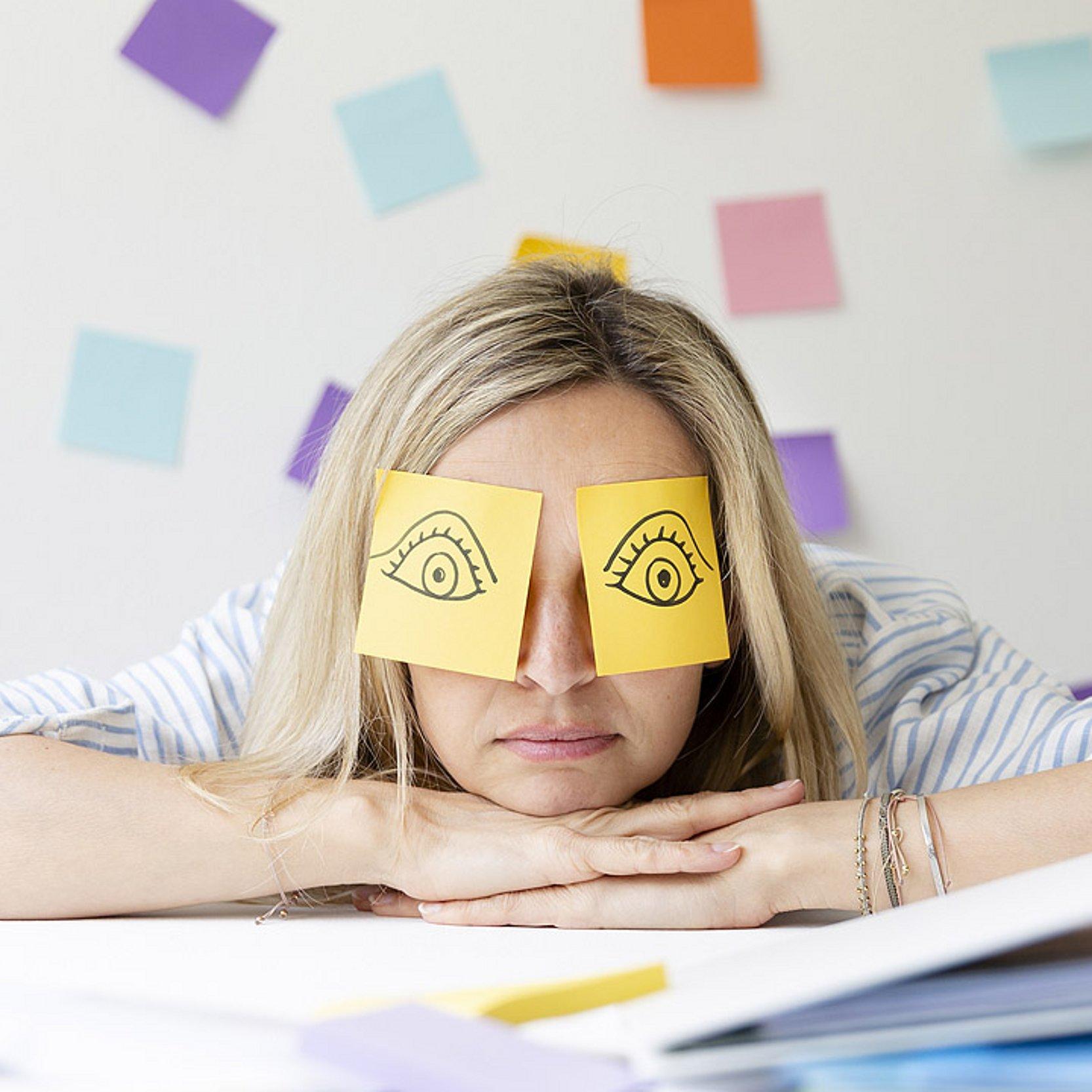 Eine Frau stützt ihren Kopf im Büro auf ihre Hände ab, auf ihren geschlossenen Augen kleben Post-It-Zettel mit aufgemalten wachen, offenen Augen