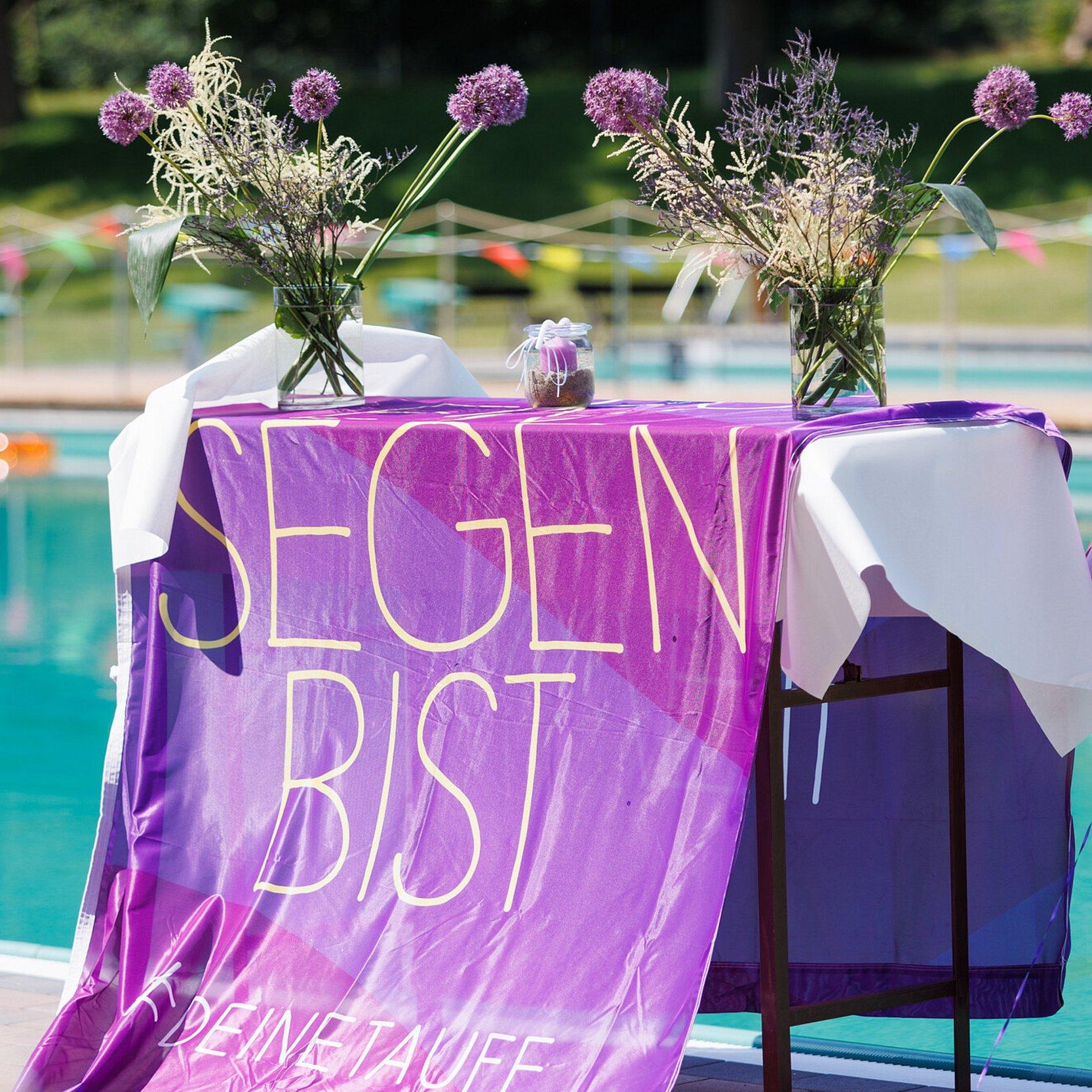 Tauffest im Naturschwimmbad Höhr-Grenzhausen: Eine Fahne der Taufinitiative der EKD liegt auf einem Tisch und hängt bis zum Boden. Der Tisch steht vor einem Schwimmbecken und ist mit Blumen und Taufkerzen dekoriert. Aufnahme seitlich von rechts.