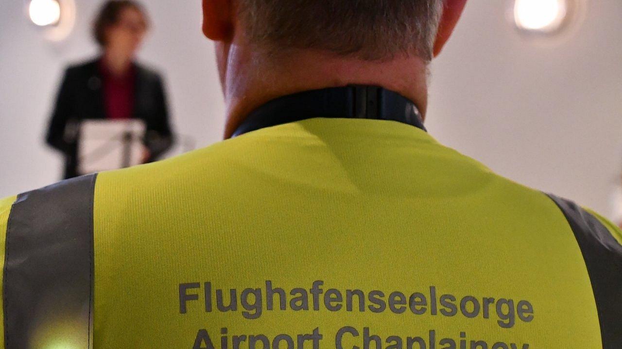 Rückansicht eines Mannes in einer gelben Warnweste mit der Aufschrift „Flughafenseelsorge Airport Chaplaincy“, im Hintergrund unscharf Kirchenpräsidentin Tietz an einem Rednerpult in einem hellen Innenraum.