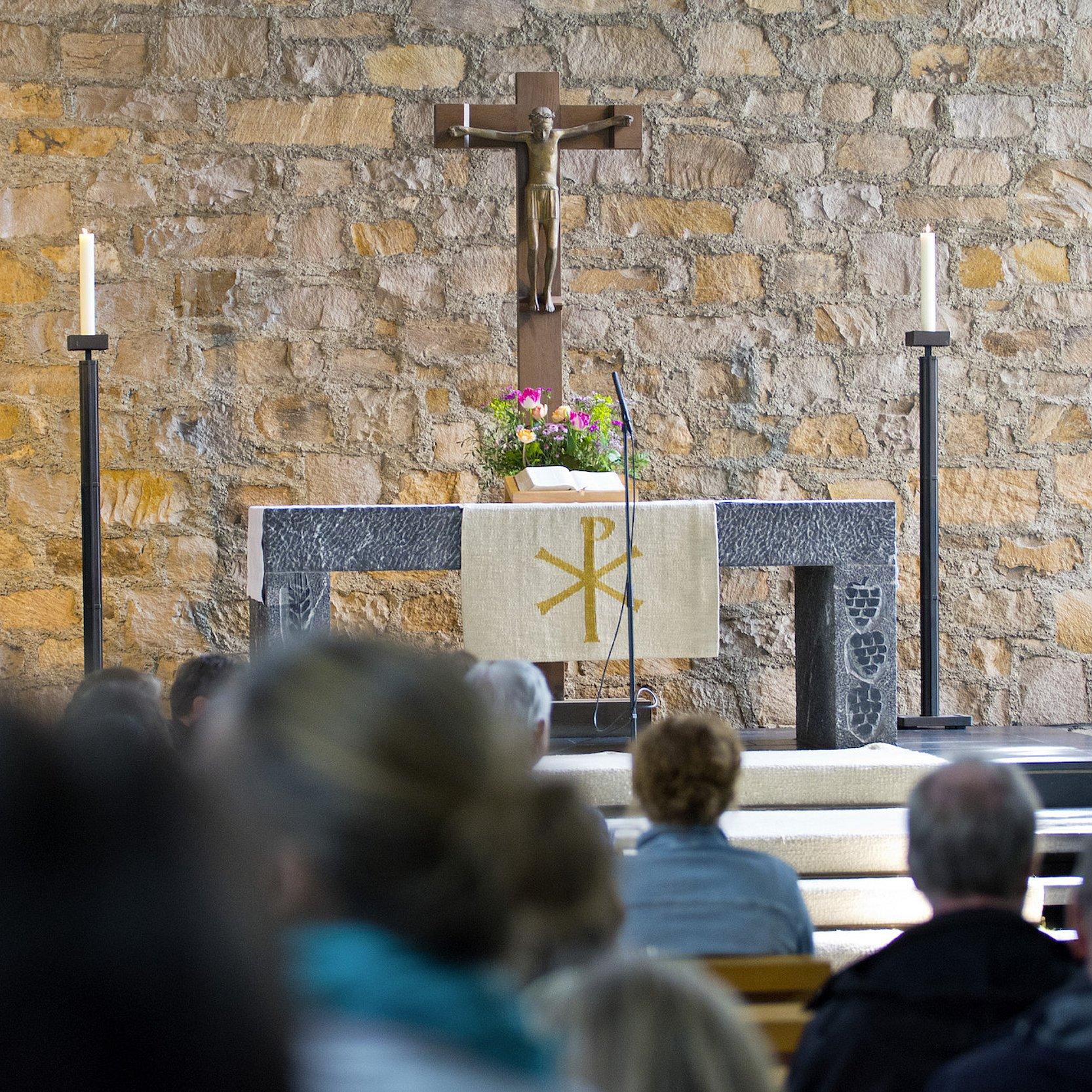 Gemeindegottesdienst in einer Kirche; im Vordergrund sitzt die versammelte Gemeinde, im Hintergrund stehen Altar, Kreuz und Kerzen vor einer Natursteinwand.
