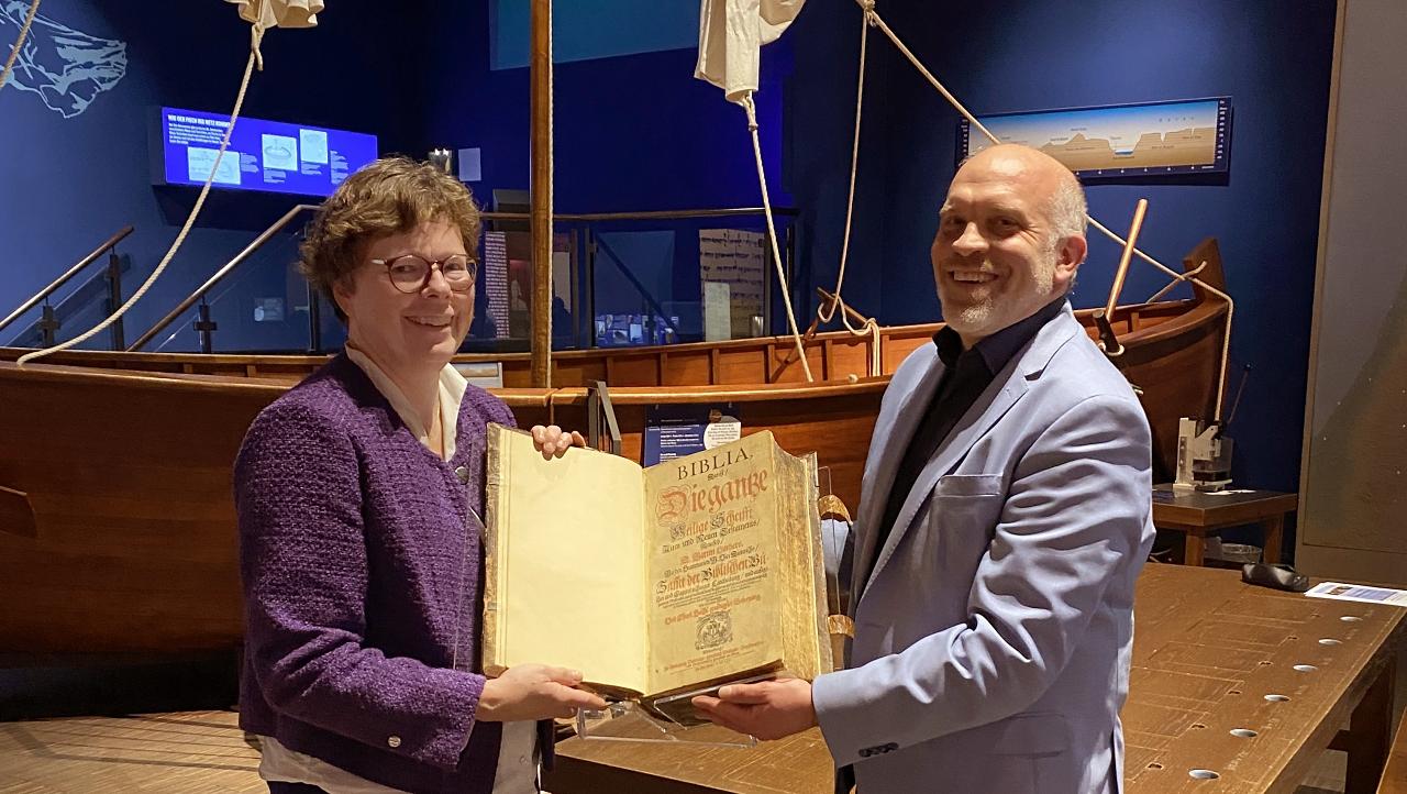Bischöfin Hofmann übergibt die historische Bibel an Museumsdirektor Veit Dinkelaker. Im Hintergrund ist ein historisches Segelschiff im Austellungsraum des Museums zu sehen.