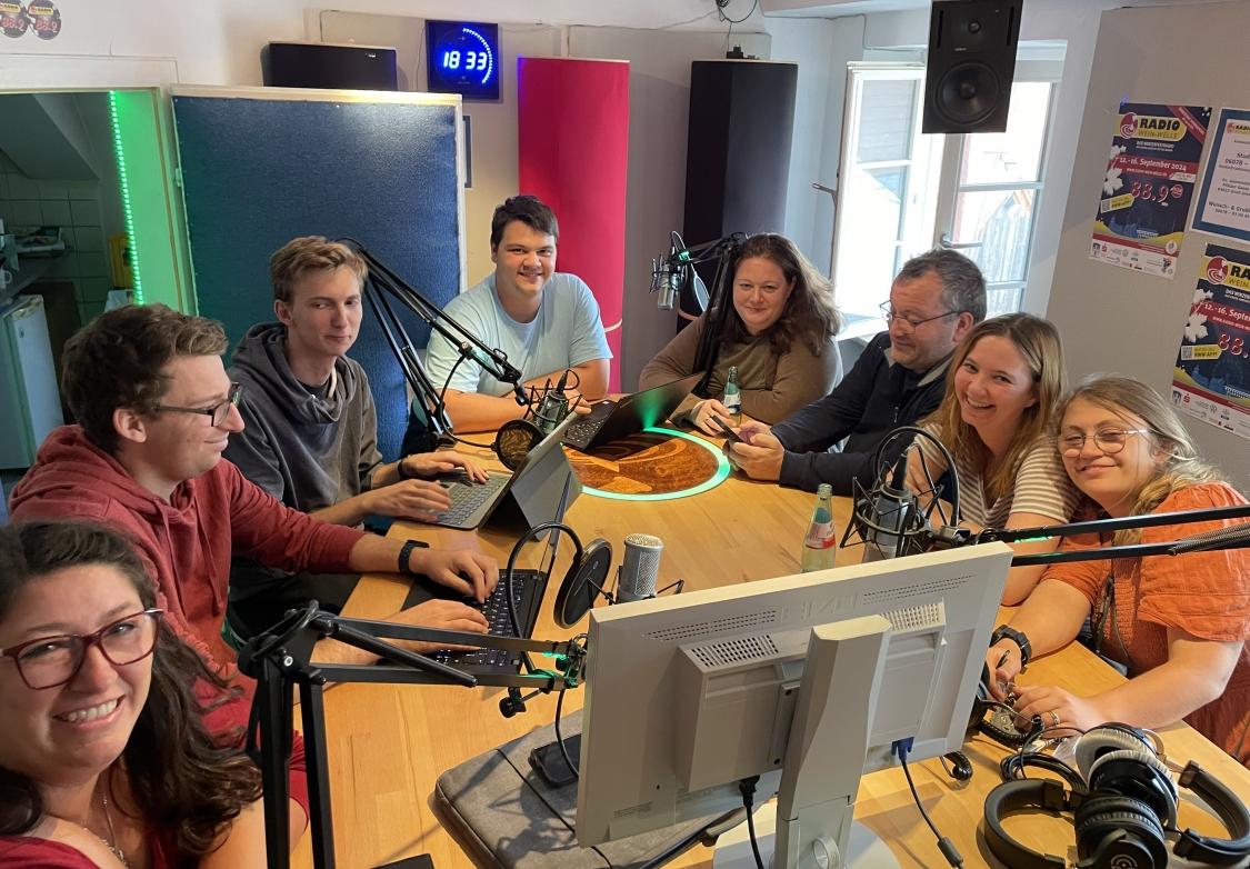 Gruppe von Personen sitzen an einem Tisch, mit Mikrofon, Kopfhörer und weiterem Studio-Equipement