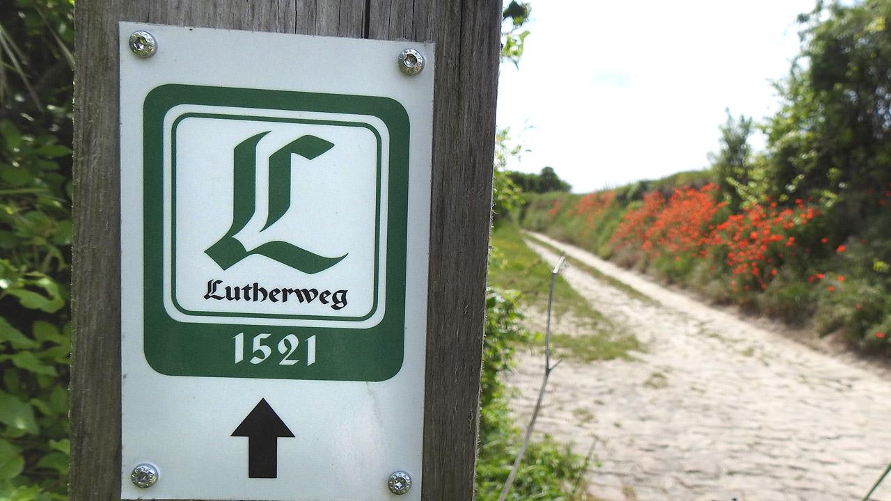 Logo des Lutherweges, daneben ein Weg, der mit roten Blumen gesäumt ist