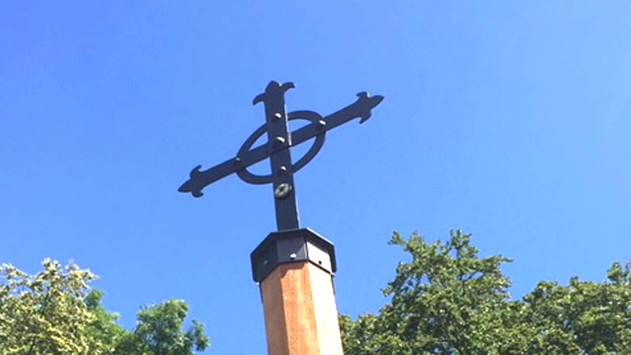 Ein schwarzes Eisenkreuz, die unmittelbare "Kreuzung" wird von einem Eisenkreuz eingerahmt, dahinter blauer Himmel