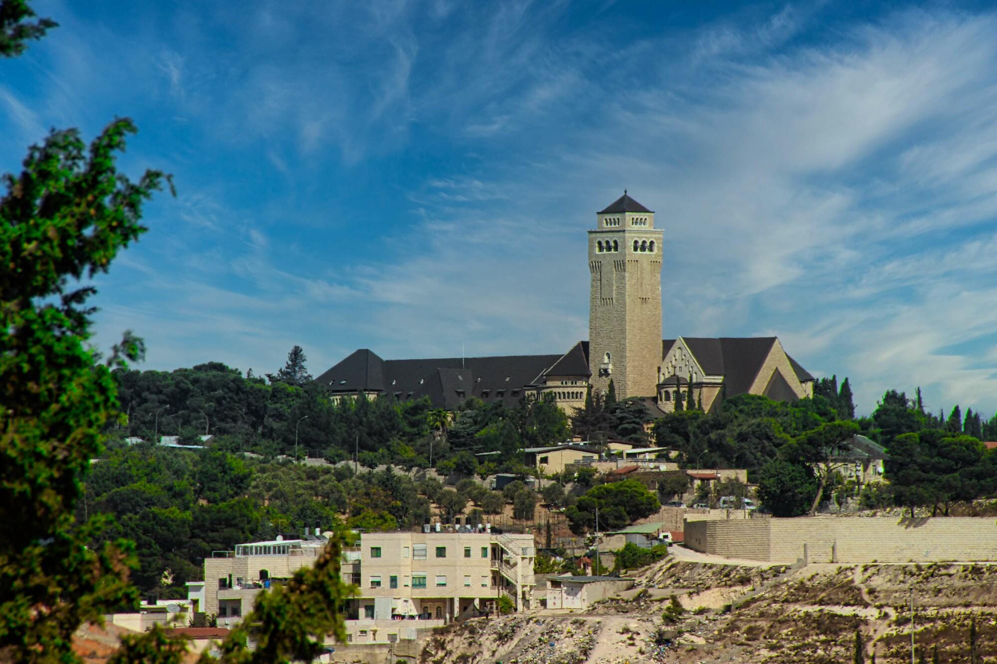 Große Kirche mit hohem Turm auf einem Hügel, umgeben von Bäumen und Wohnhäusern, unter einem blauen Himmel mit feinen Wolken.