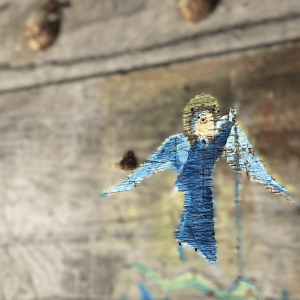 Ein gemalter Engel mit Heiligenschein, dunkelblauem Gewand und hellblauen Flügeln auf einer Holztüre. Die Holztüre im Hintergrund ist unscharf, so dass der Engel deutlich hervortritt