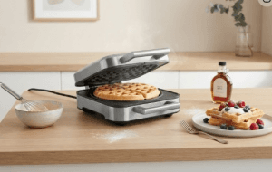 Ein Waffeleisen steht auf einem Tisch. Daneben ein Teller mit zwei Waffeln, die mit Beeren belegt sind.