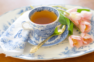 Eine gefüllte Teetasse steht auf einem Kuchenteller, daneben liegt ein goldfarbener Teelöffel.