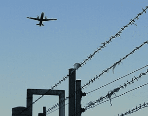 Flugzeug am Himmel, darunter ein Stacheldrahtzaun