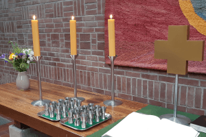 Altar der Johannesgemeinde, bunter Blumenstrauß, 3 brennende Kerzen, davor 2 Tablets mit Einzelkelchen für das Abendmahl, daneben das Kreuz und davor die aufgeschlagene Bibel