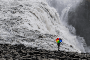 Das Bild zeigt das Schwarzweißfoto eines Wasserfalls. Davor steht ein Mensch mit dem Rücken zur Kamera und einem riesigen Sonnenschirm in Regenbogenfarben.