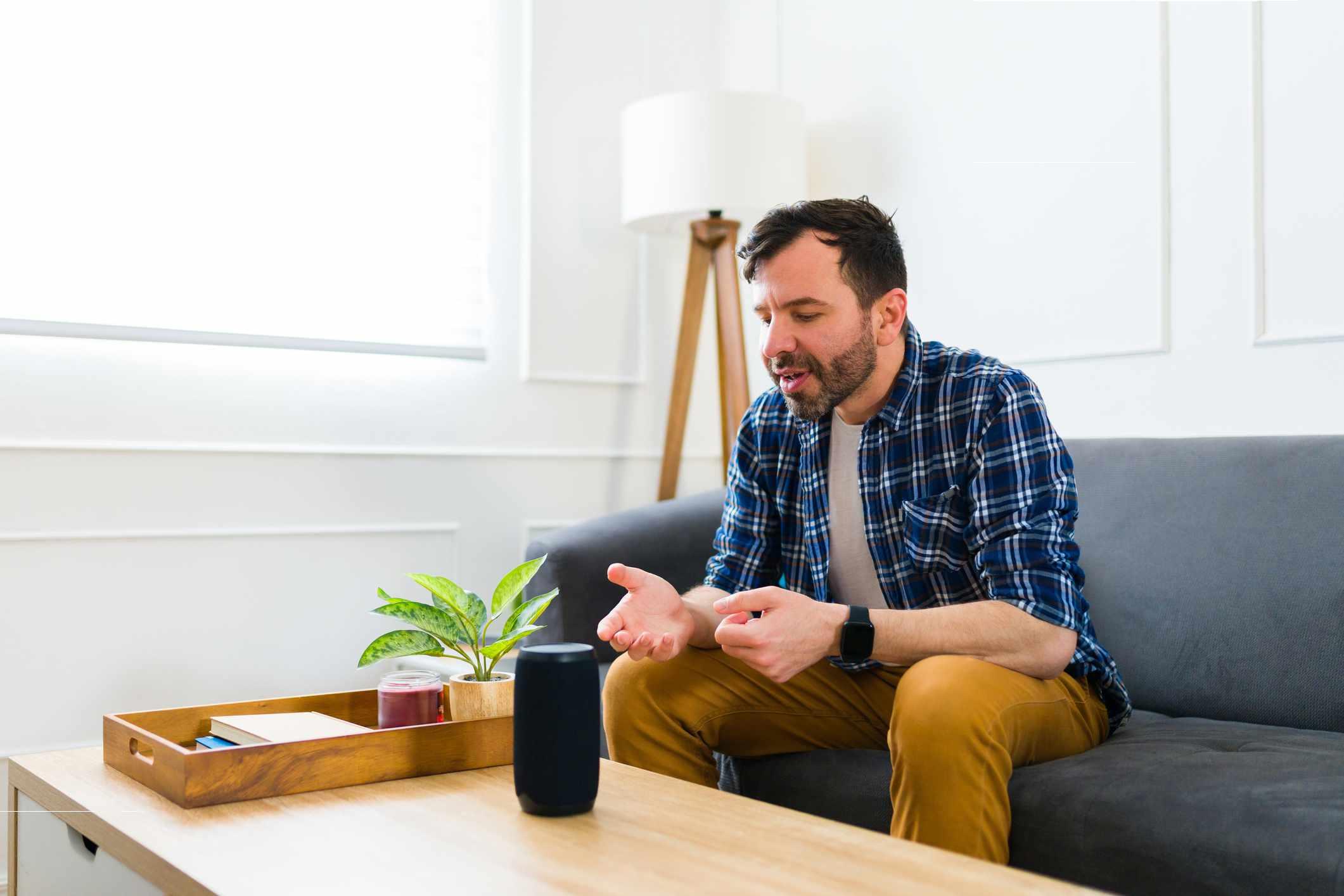 Ein Mann sitzt auf dem Sofa und Sprachassistentin Alexa steht auf dem Tisch vor ihm. Der Mann spricht zu Alexa.