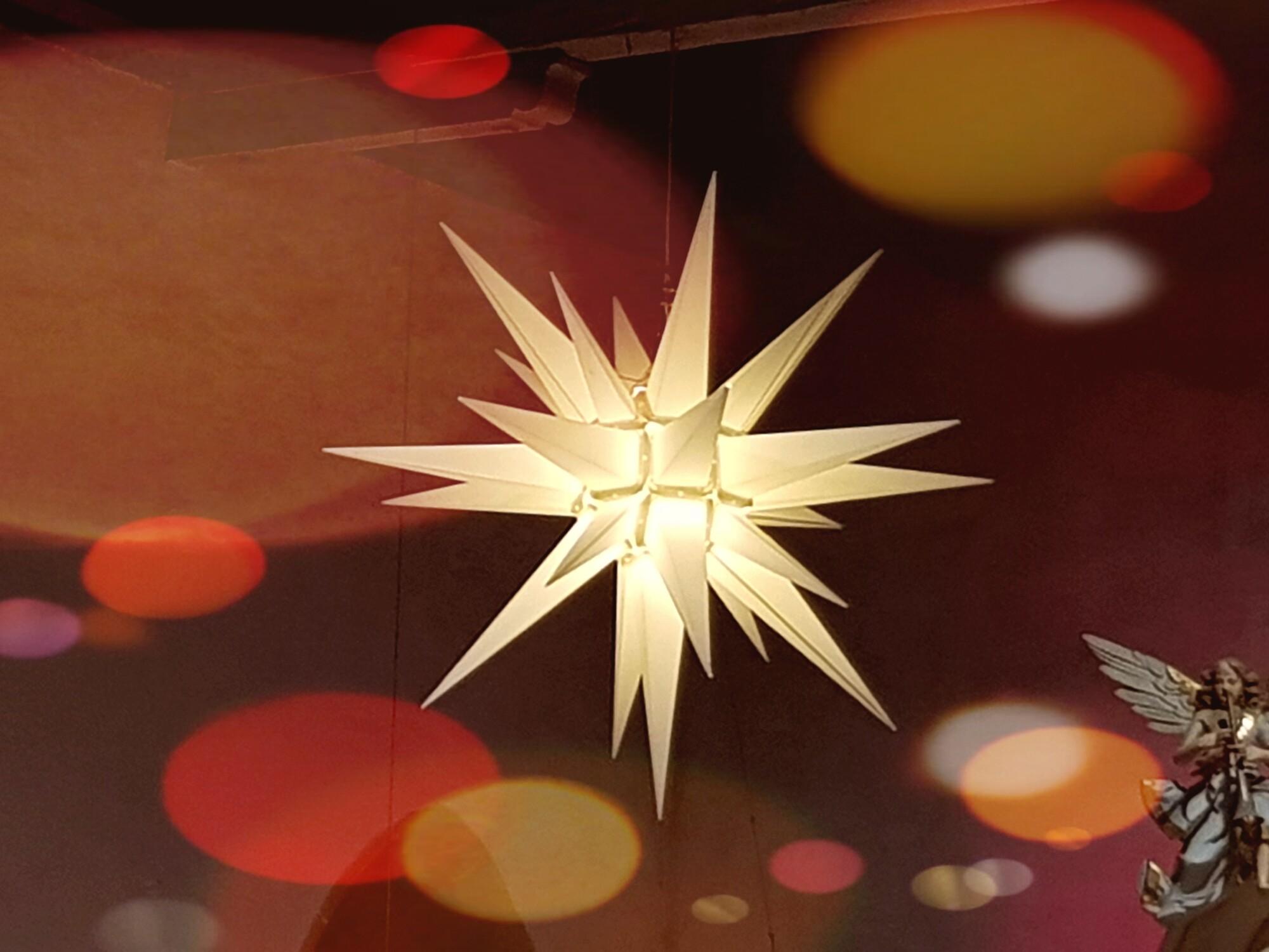 Herrnhuter Stern in der St.-Marien-Kirche Bleicherode Das Bild zeigt einen weißen Herrnhuter Stern mit bunten Lichtpunkten an der Kirchendecke. In der rechten unteren Ecke ist ein Engel zu sehen.