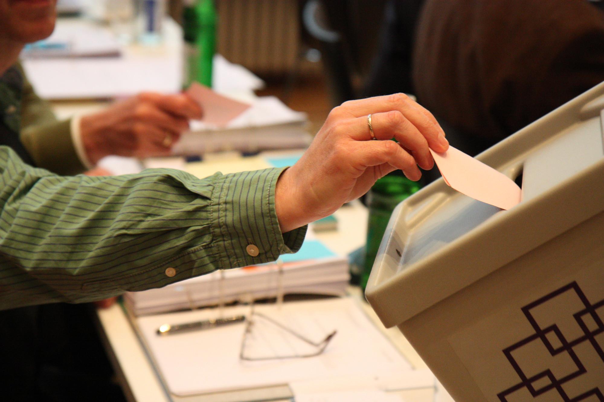 Eine Hand steckt einen Wahlzettel in eine Wahlurne mit einem Logo der EKHN..