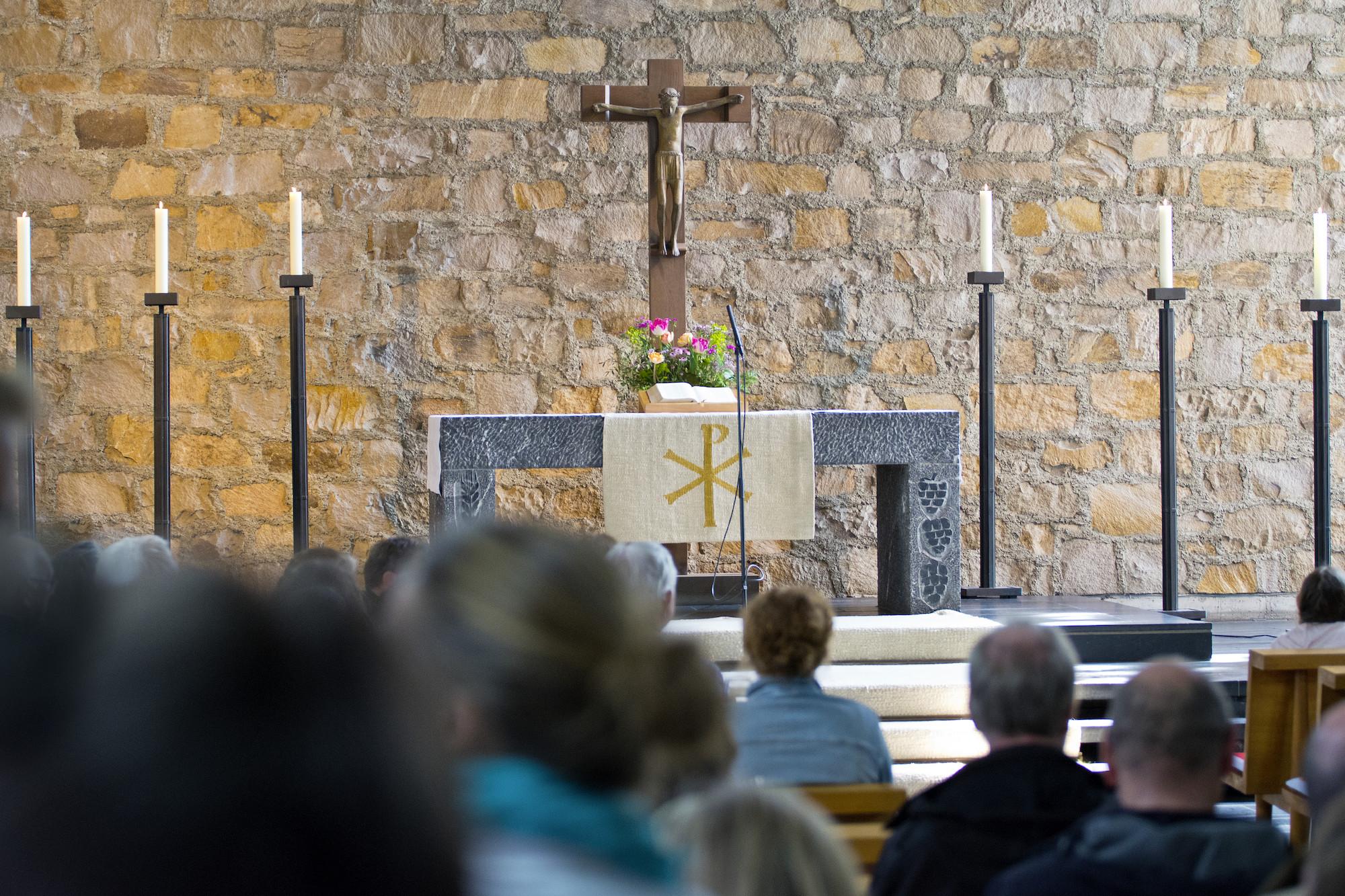 Kirchenraum Altar Gemeindegottesdienst in einer Kirche; im Vordergrund sitzt die versammelte Gemeinde, im Hintergrund stehen Altar, Kreuz und Kerzen vor einer Natursteinwand.