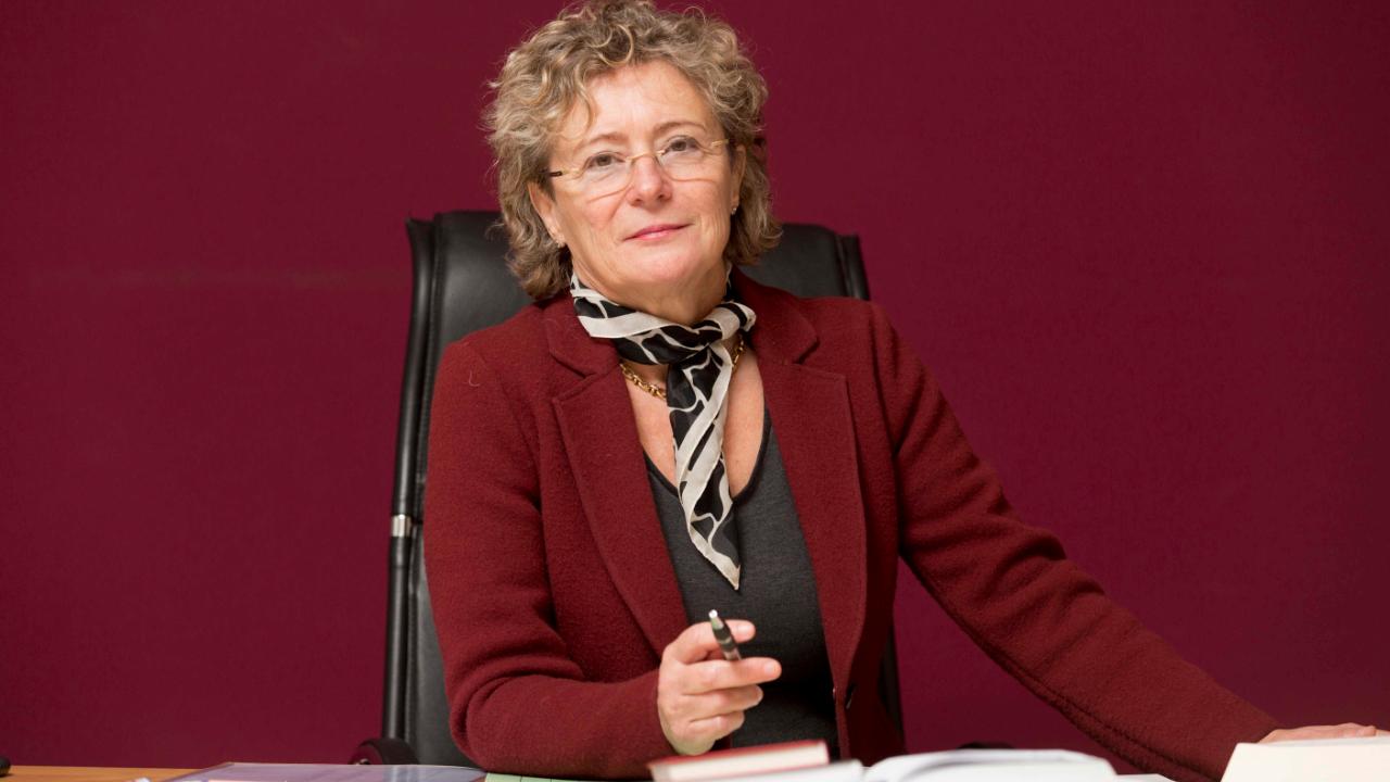 Esther Gebhardt Esther Gebhardt mit rotem Blazer und buntem Halstuch sitzt an ihrem Schreibtisch und hält einen Kugelschreiber in der Hand