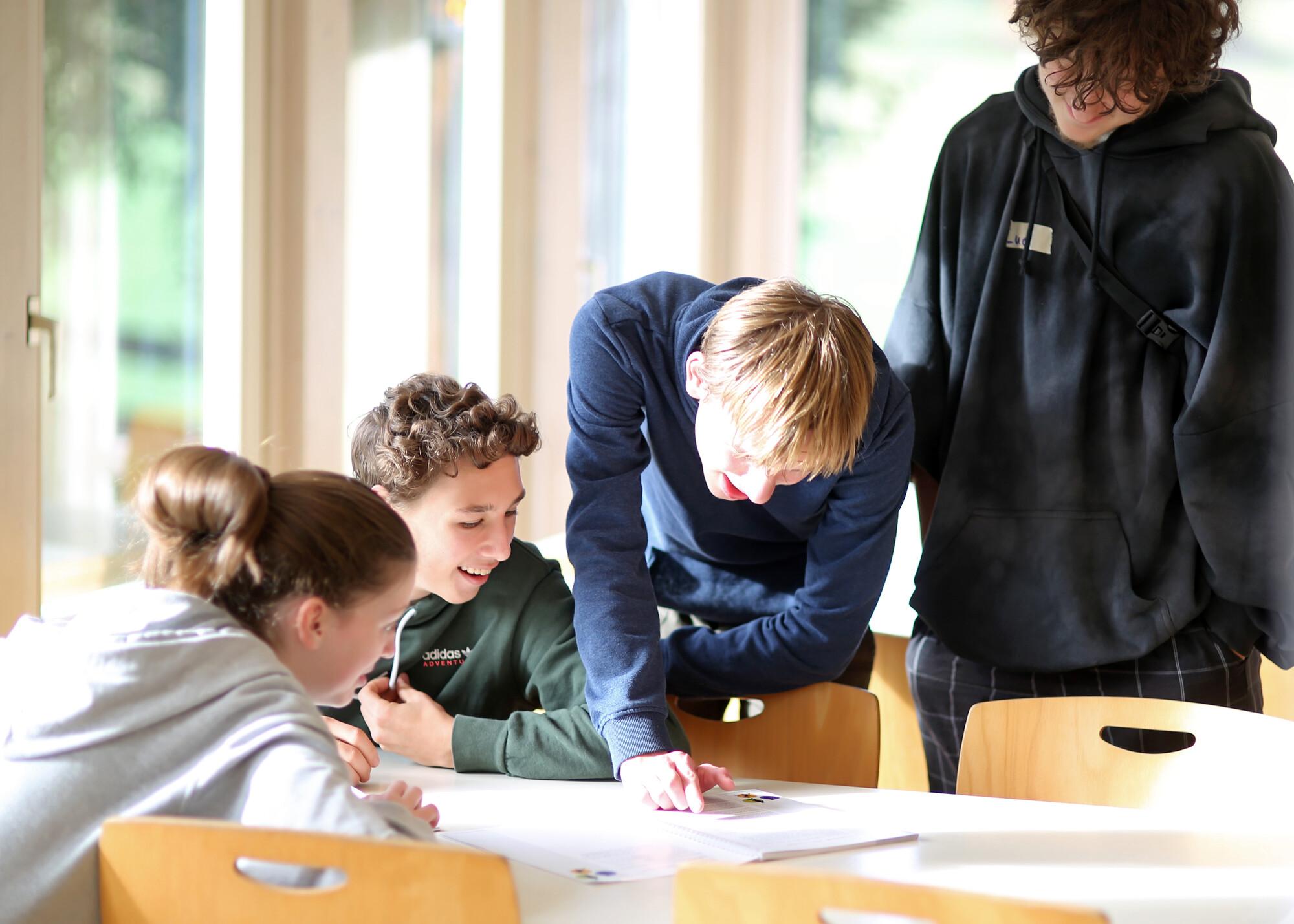 Jugendliche bei Besprechung Links sitzen zwei Jugendliche an einem Tisch, rechts daneben über den Tisch gebeugt und stehend sind zwei andere Jugendliche zu sehen. Sie reden freundlich miteinander.