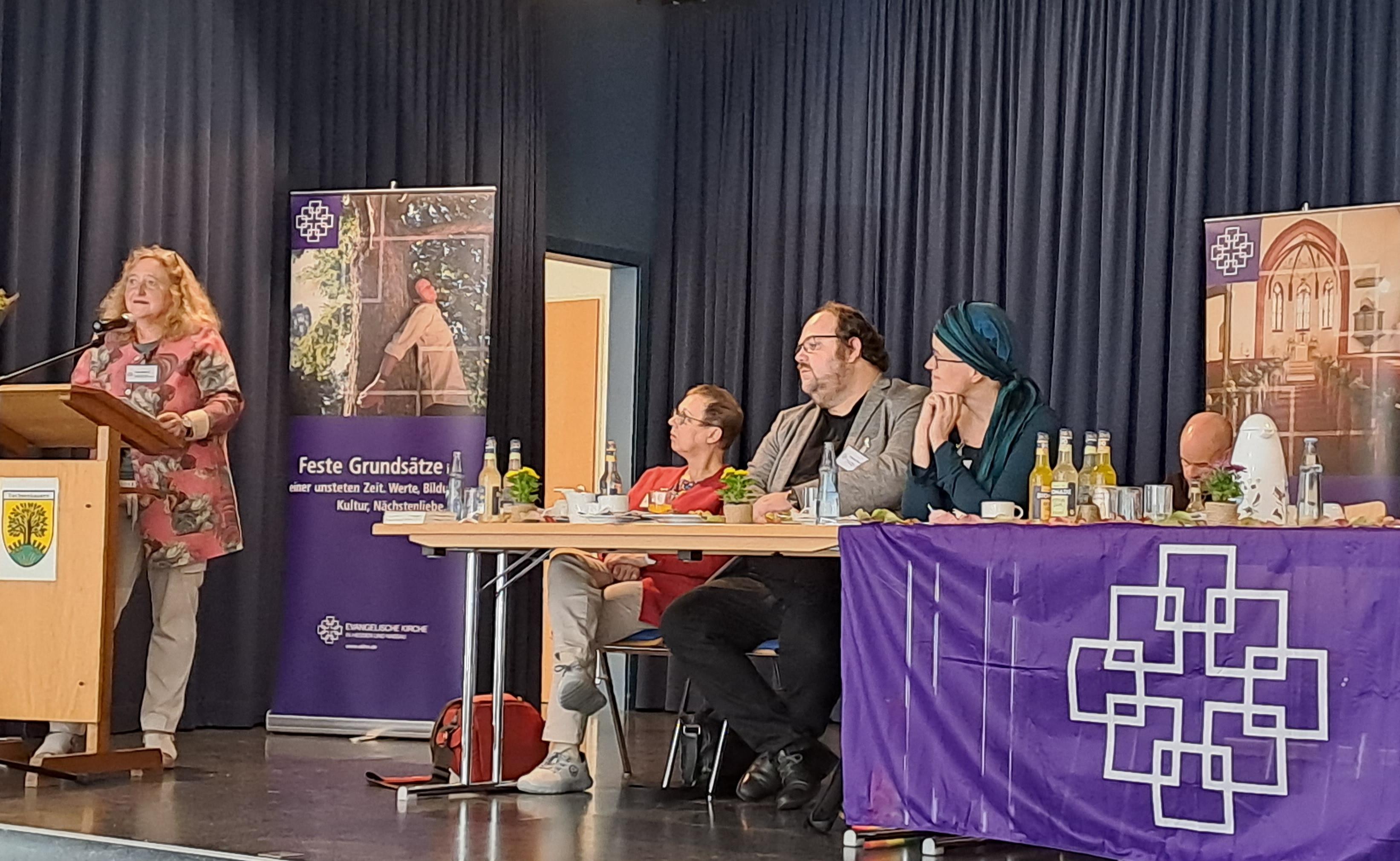 Auf einem Podium steht links eine Frau am Rednerpult. Rechts sitzen mehrere Personen hinter einem Tisch Das Podium ist geschmückt mit lila Banner der Evangelischen Kirche in Hessen und Nassau.