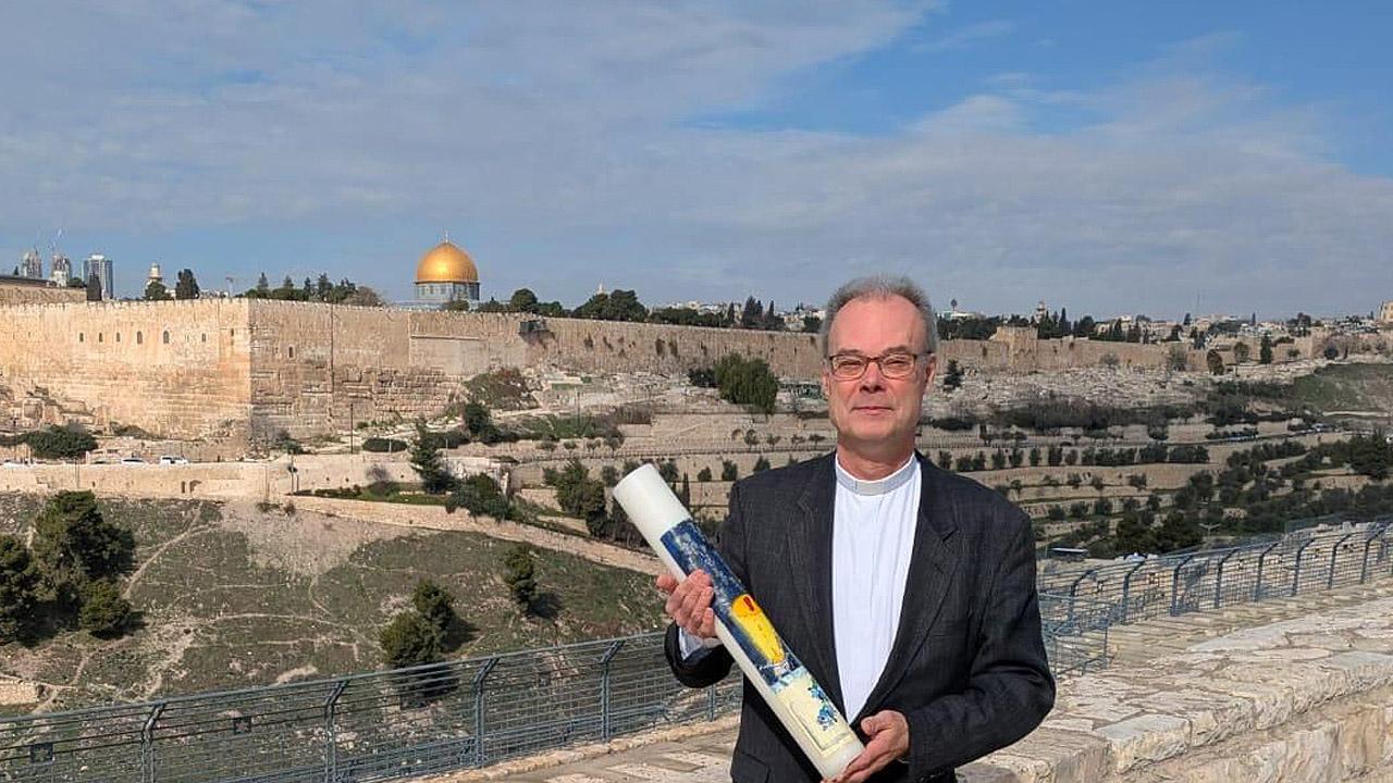 Andreas Goetze in Jerusalem Ein Mann im dunklen Anzug trägt eine große Kerze vor der Stadtansicht Jerusalems mit Felsendom