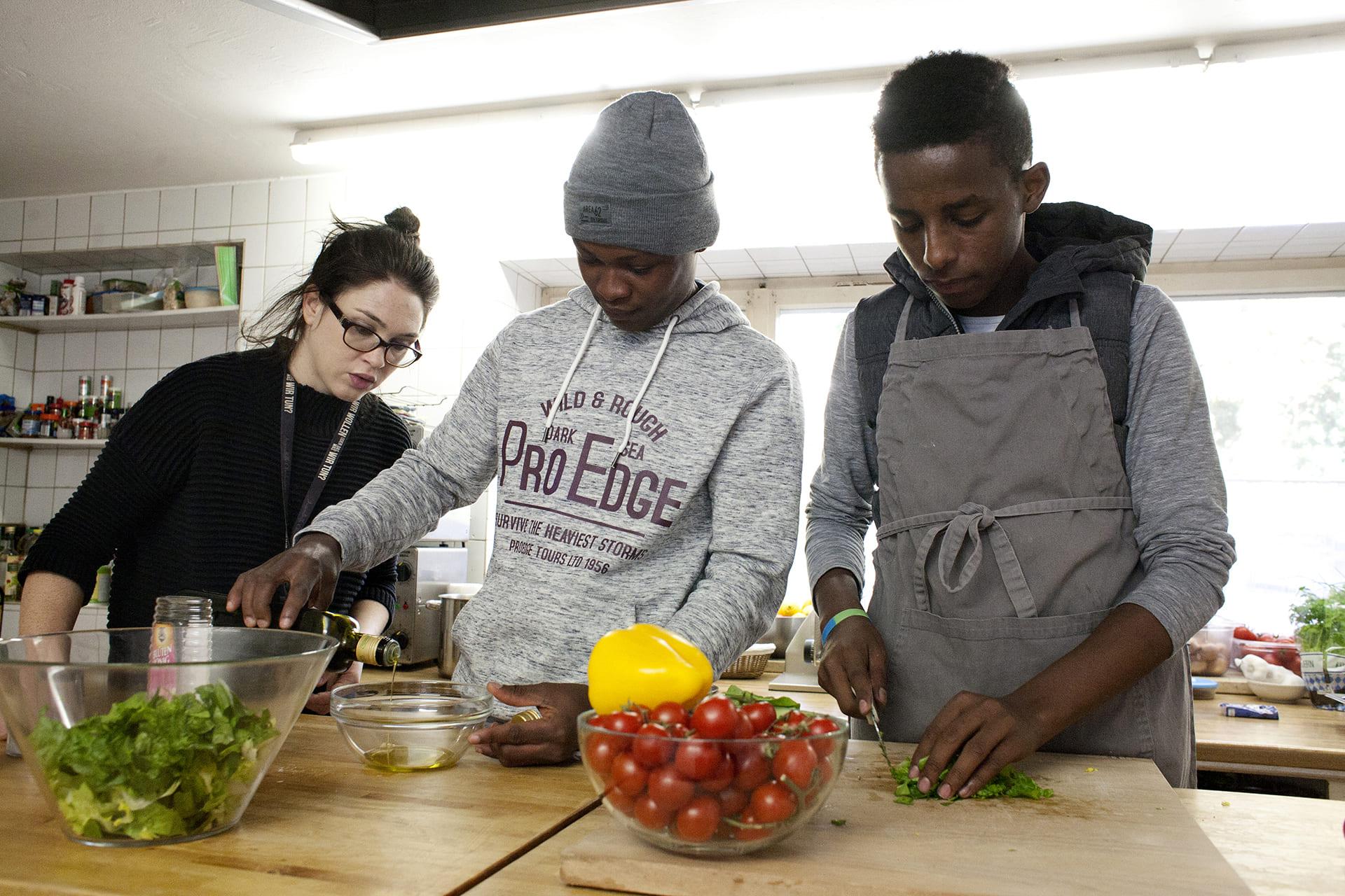 Zwei junge Geflüchtete kochen gemeinsam mit einer jungen Frau im Vordergrund steht eine Schüssel mit Tomaten.