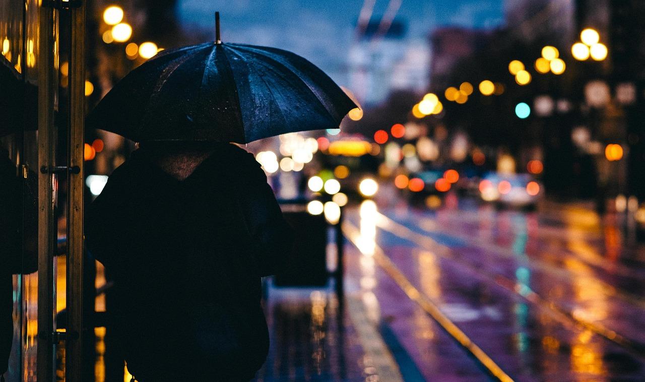 Einsam in der Nacht „Eine Person mit Regenschirm steht bei Regen an einer beleuchteten Straße in der Dämmerung. Das unscharfe Stadtlicht erzeugt eine nachdenkliche und ruhige Atmosphäre.“