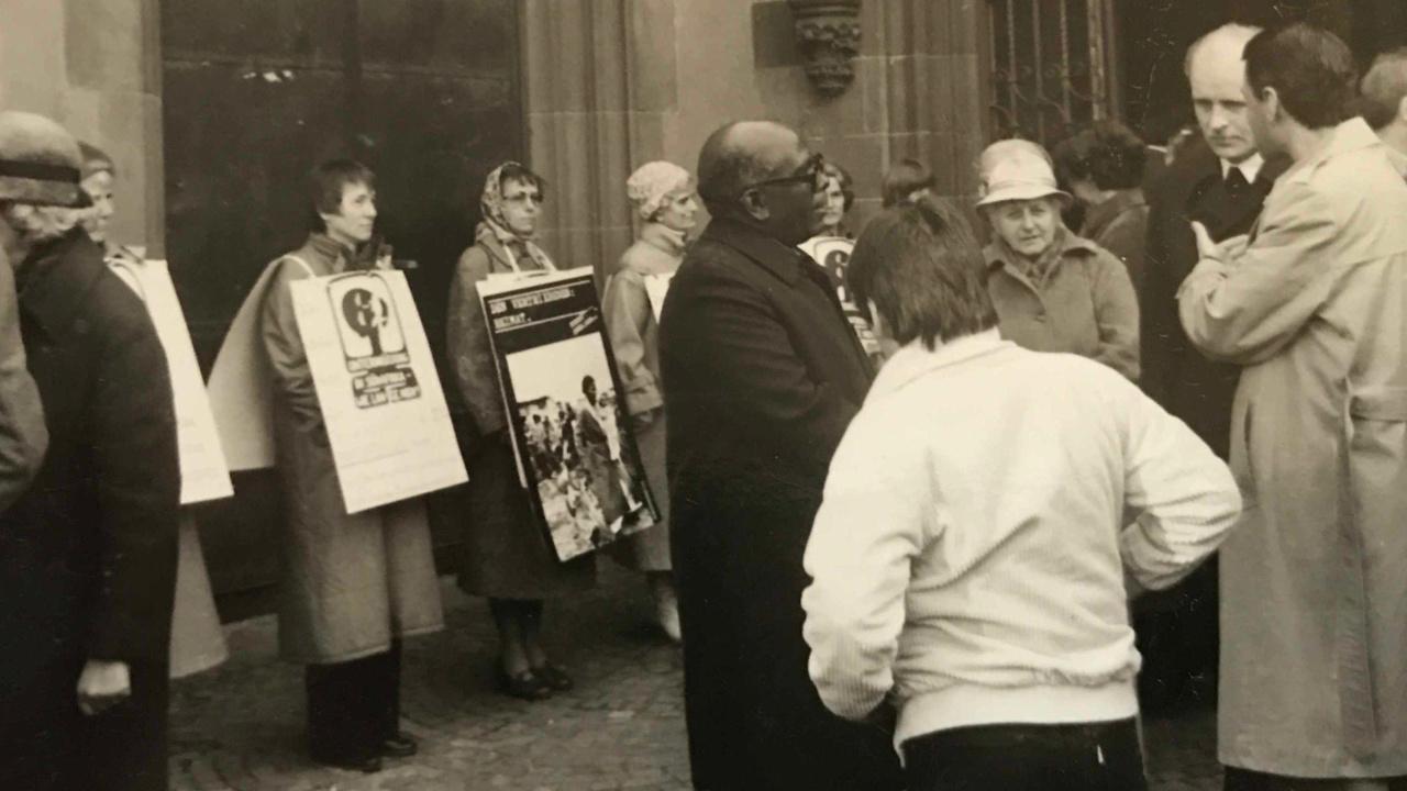 Frauen für Südafrika - Mahnwache vor dem Frankfurter Dom Frauen stehen mit Plakaten, die sie umgehängt haben vor dem Portal des Frankfurter Doms. Im Vordergrund steht eine Gruppe von Menschen und diskutert miteinander.